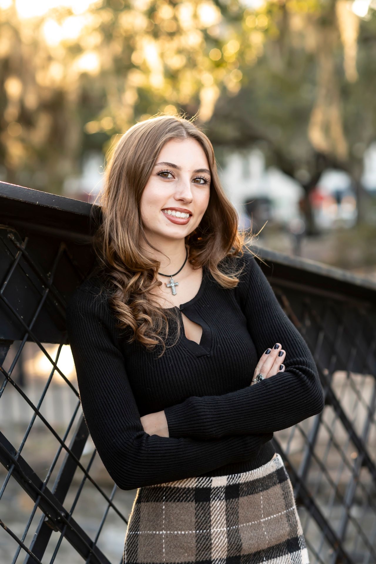 A person in a black sweater and plaid skirt stands with crossed arms on a bridge during golden hour.