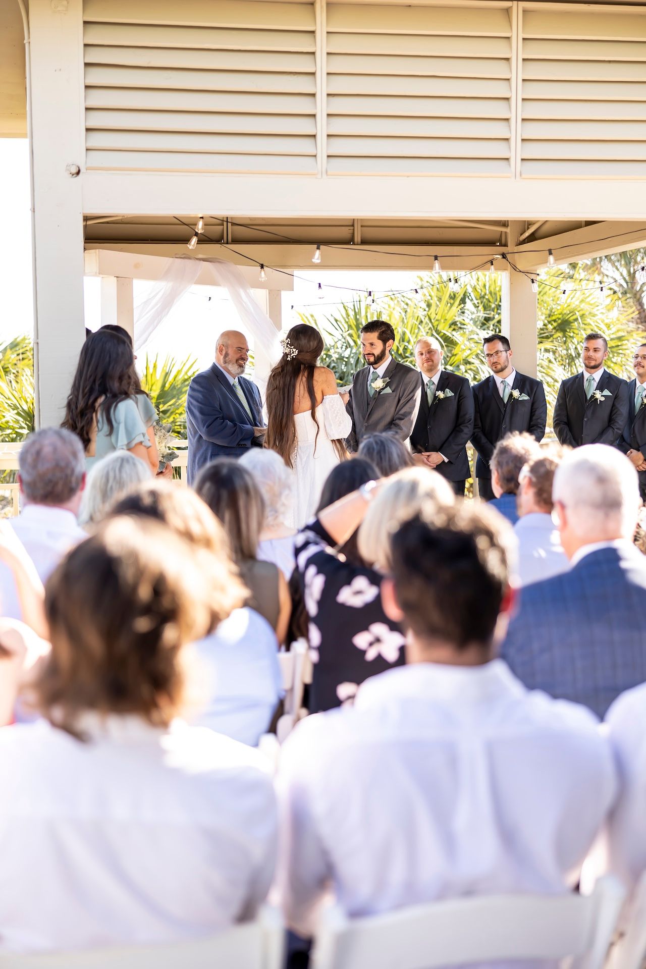 An outdoor wedding ceremony takes place under a covered pavilion surrounded by seated guests on a sunny day.