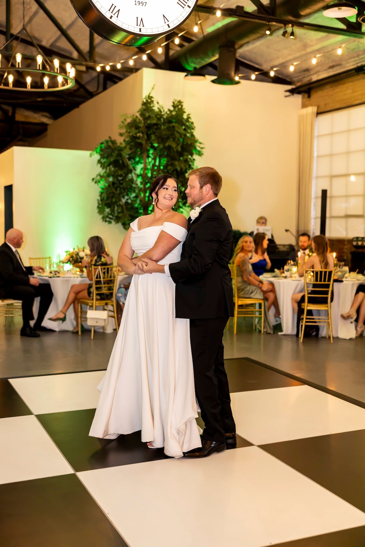 Couple shares romantic first dance on black and white checkered dance floor at elegant wedding reception with green uplighting.