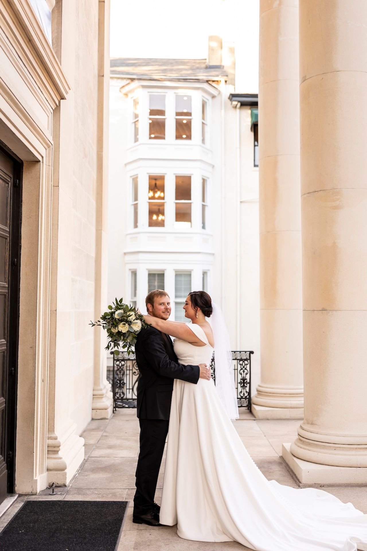 A couple in wedding attire embrace between elegant white columns of a historic building with a trailing bridal gown.