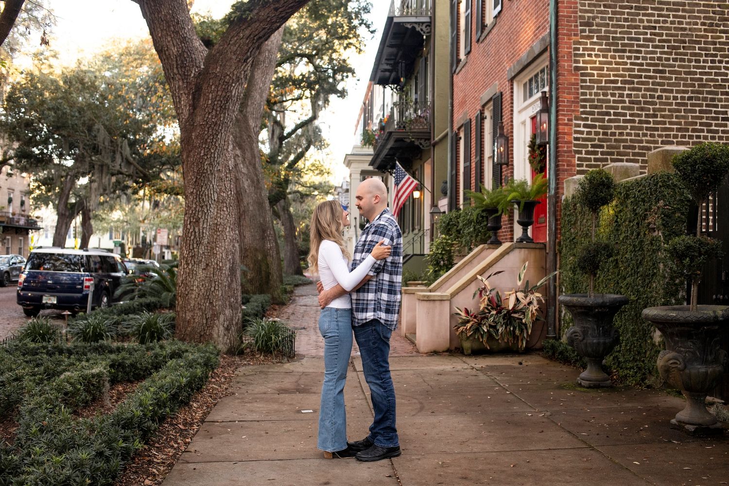 A couple embraces on a tree-lined residential street with brick row houses and parked cars in the background.