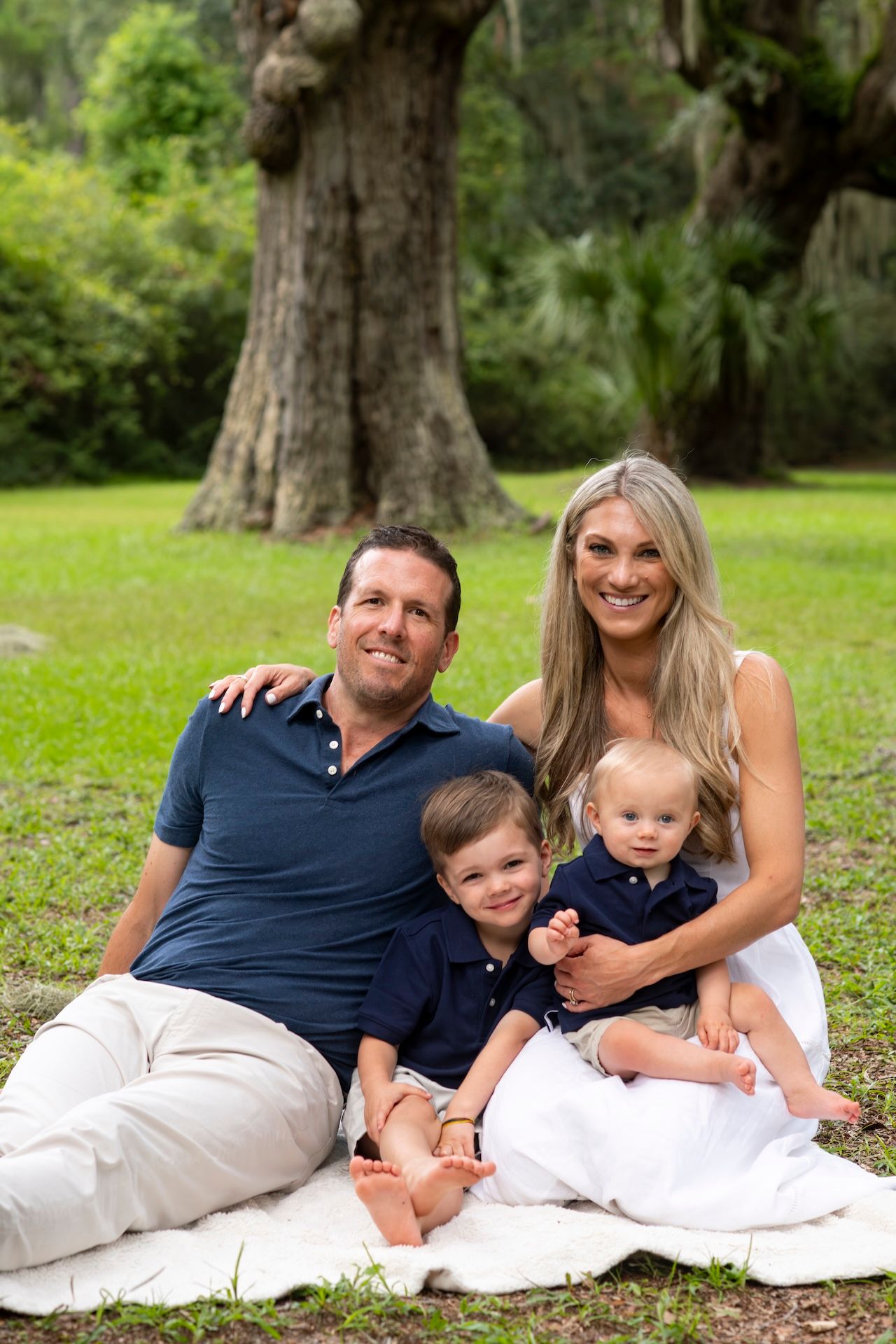 Happy family of four sitting on a blanket in a lush green park, parents smiling with two young boys.