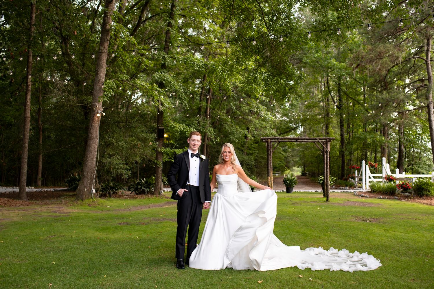 Bride and groom posing outdoors on lush green lawn surrounded by tall trees at elegant wedding venue.