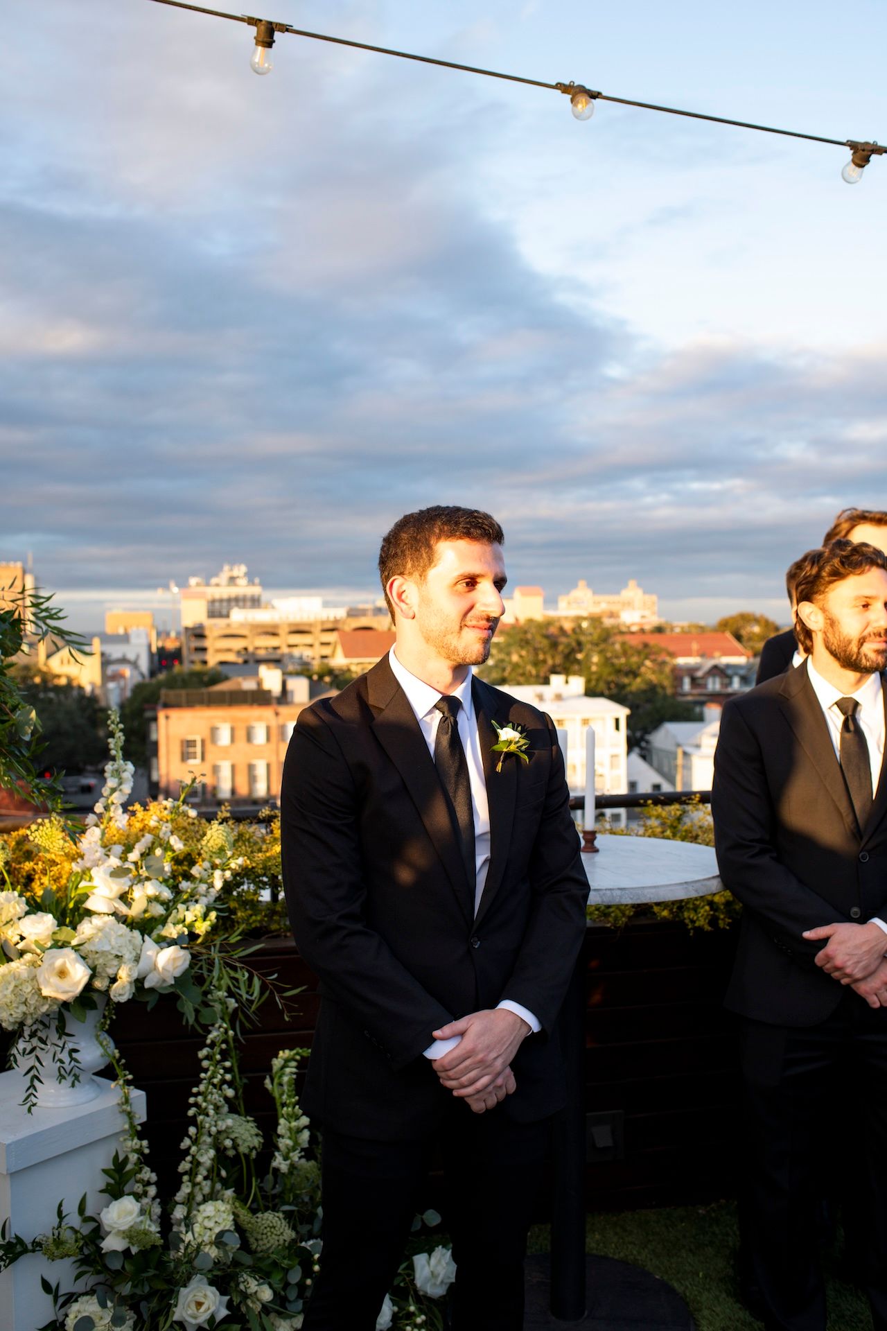 Wedding party members in dark suits stand together on a rooftop terrace at sunset with white floral arrangements.