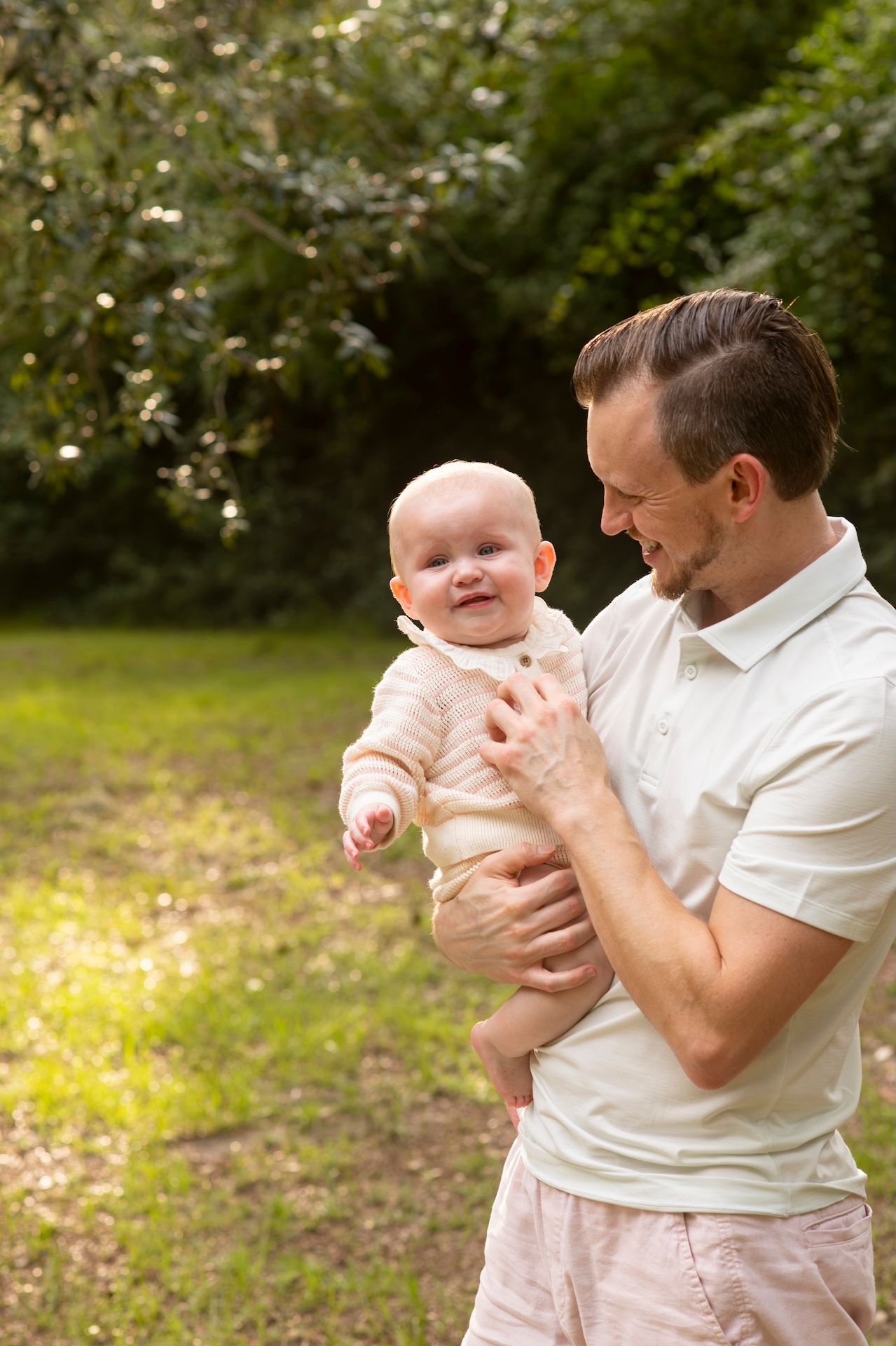 A smiling father holds his happy baby outdoors in a sunlit green park setting.