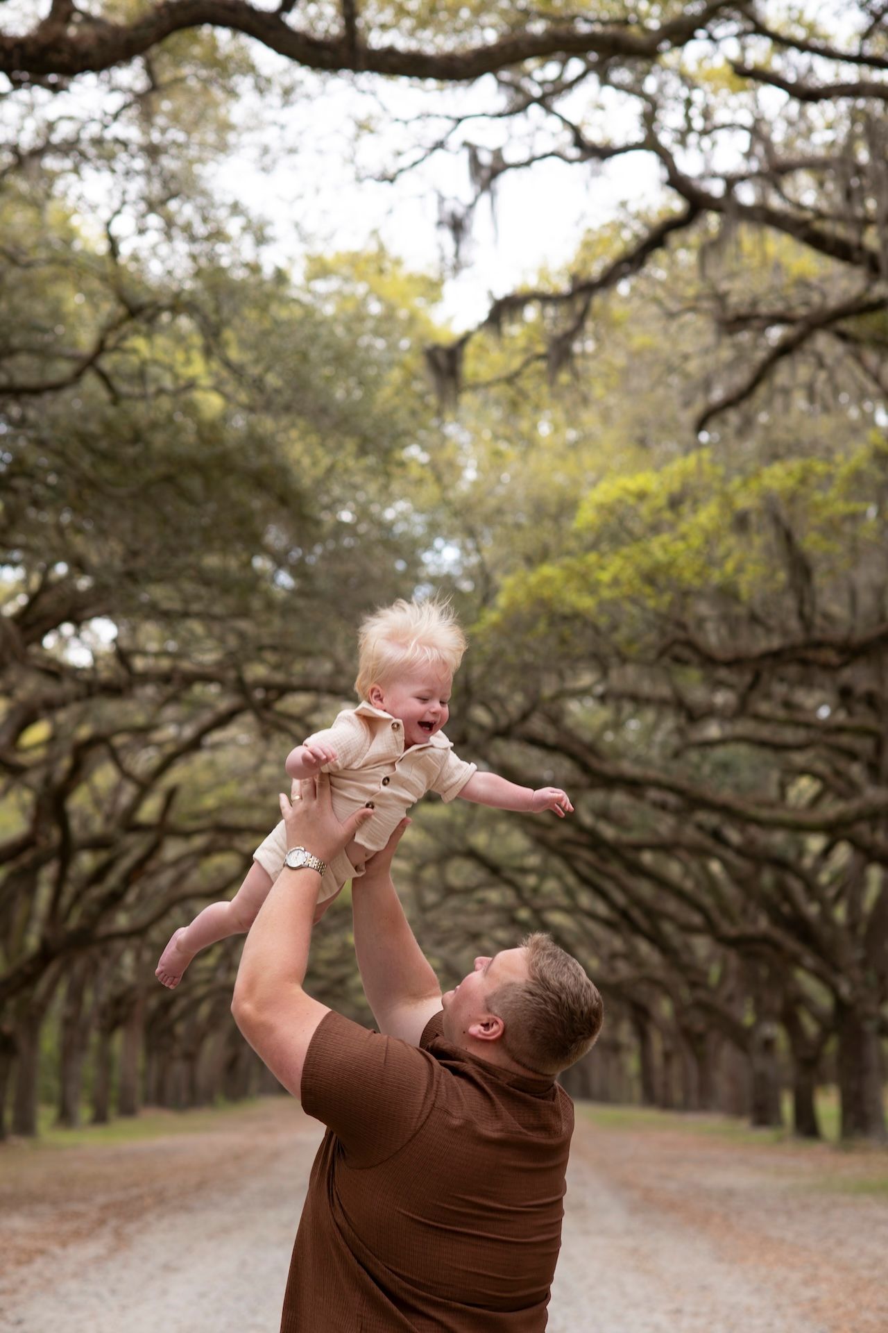 Father joyfully lifting laughing baby high in the air on a scenic tree-lined dirt path.