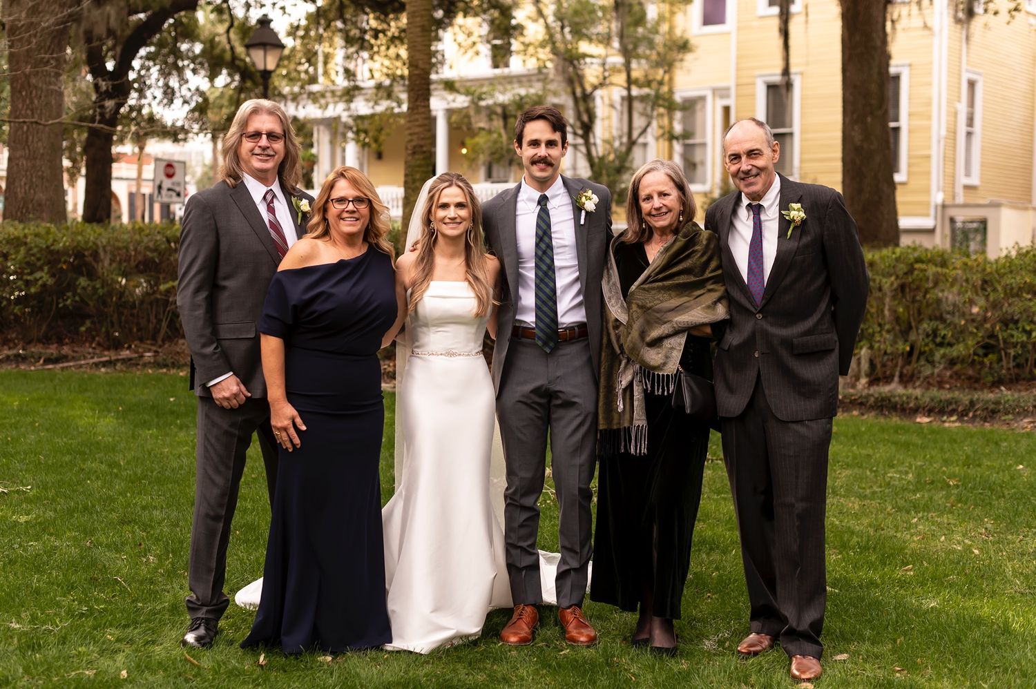 A group of formally dressed wedding guests pose together on a grassy lawn near a building in autumn.