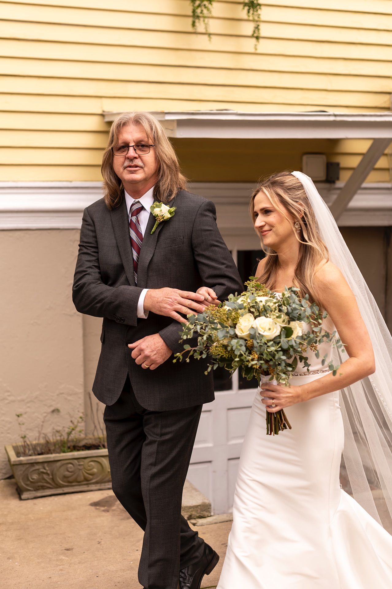 A wedding scene unfolds against a yellow building with white trim as attendees gather for a ceremony.