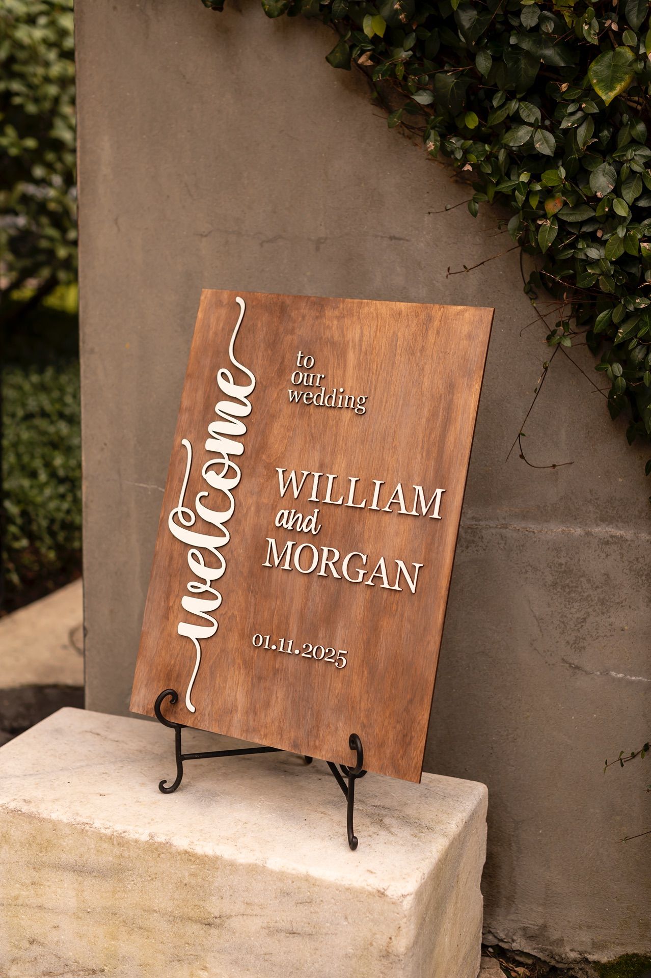 Wooden wedding signs with engraved names Morgan and William displayed on easels against a concrete wall with ivy background.