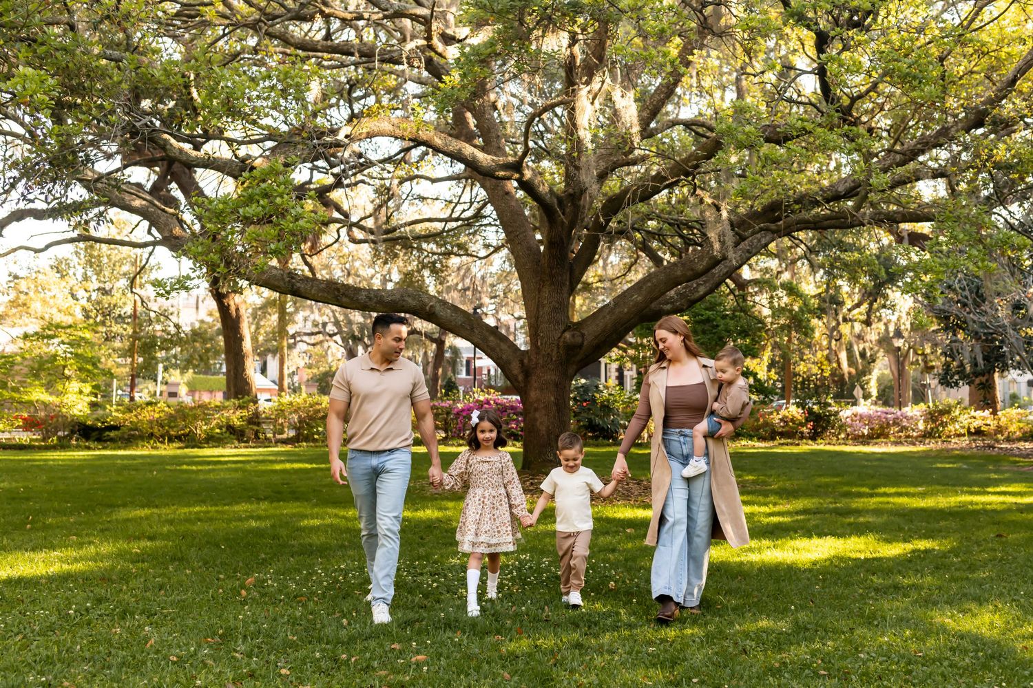 Family walking together under a large oak tree in a sunlit park setting.