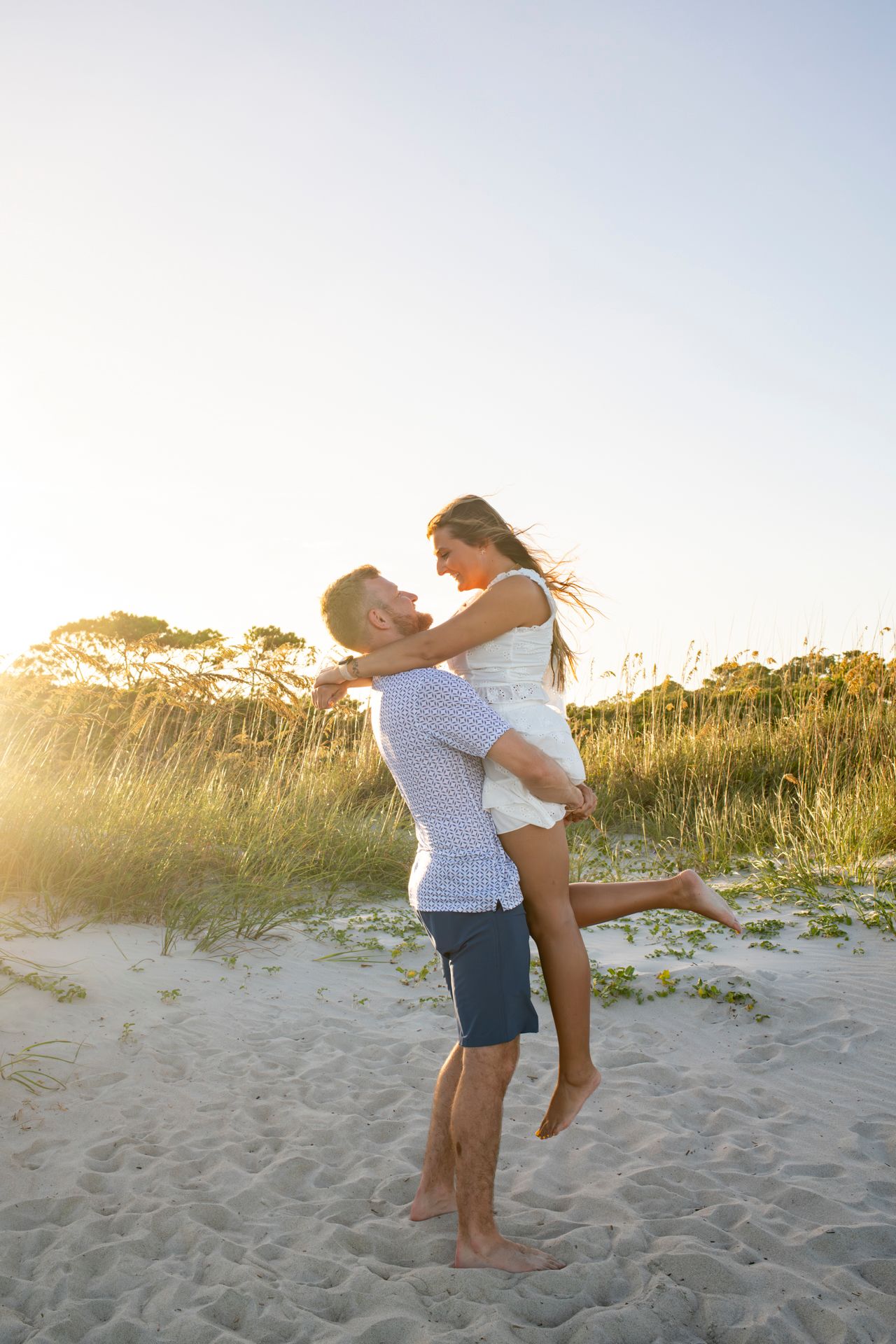 A romantic couple embraces and spins together on a sandy beach at sunset with tall grass in the background.