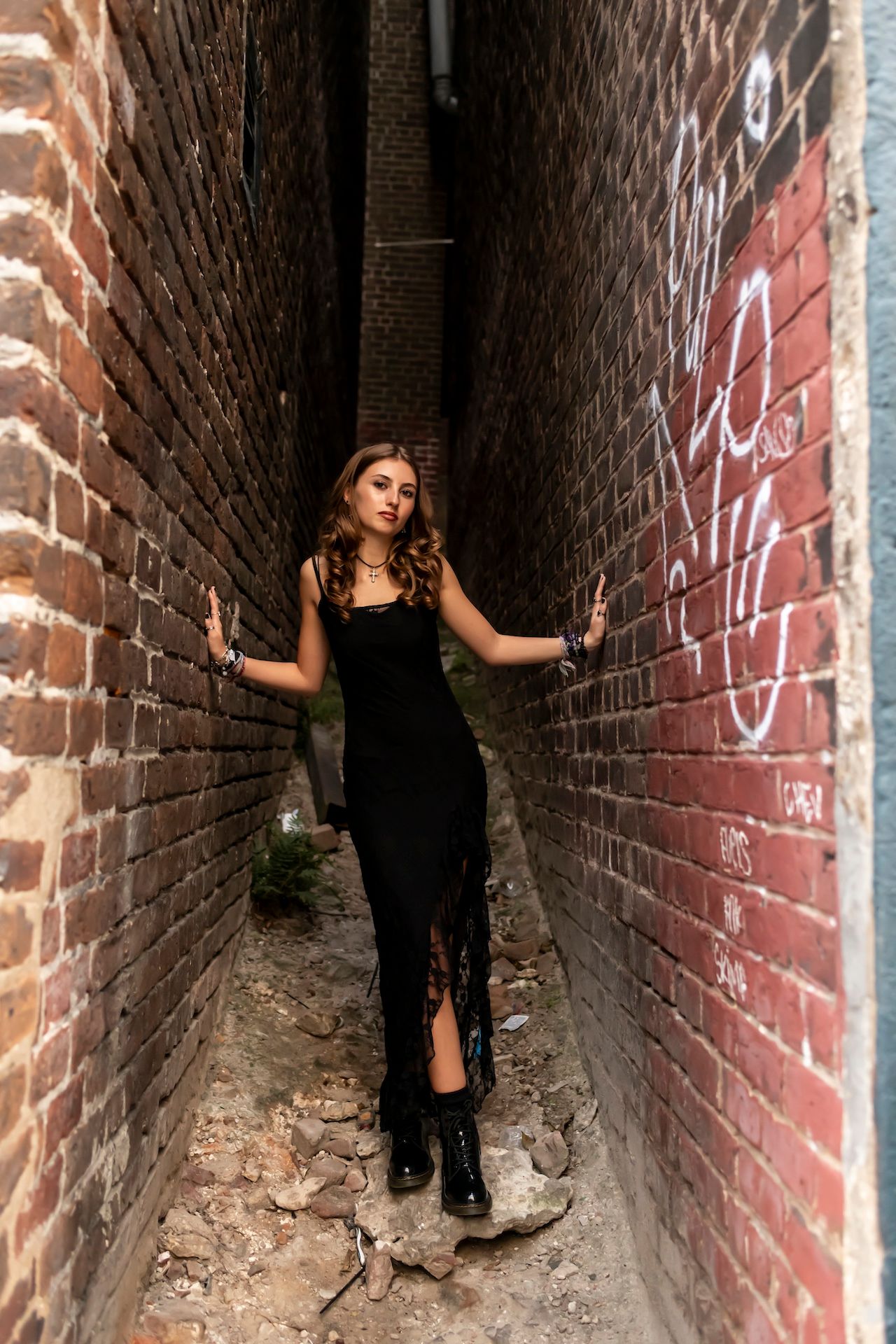 A person in a black dress poses dramatically in a narrow brick alleyway with graffiti on the walls.