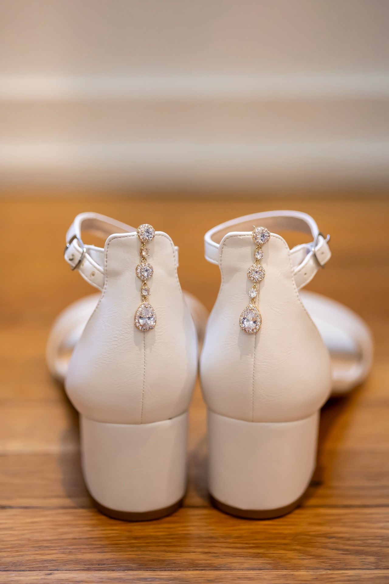 Close-up view of elegant white bridal shoes with crystal T-strap detail and low block heels on wooden surface.