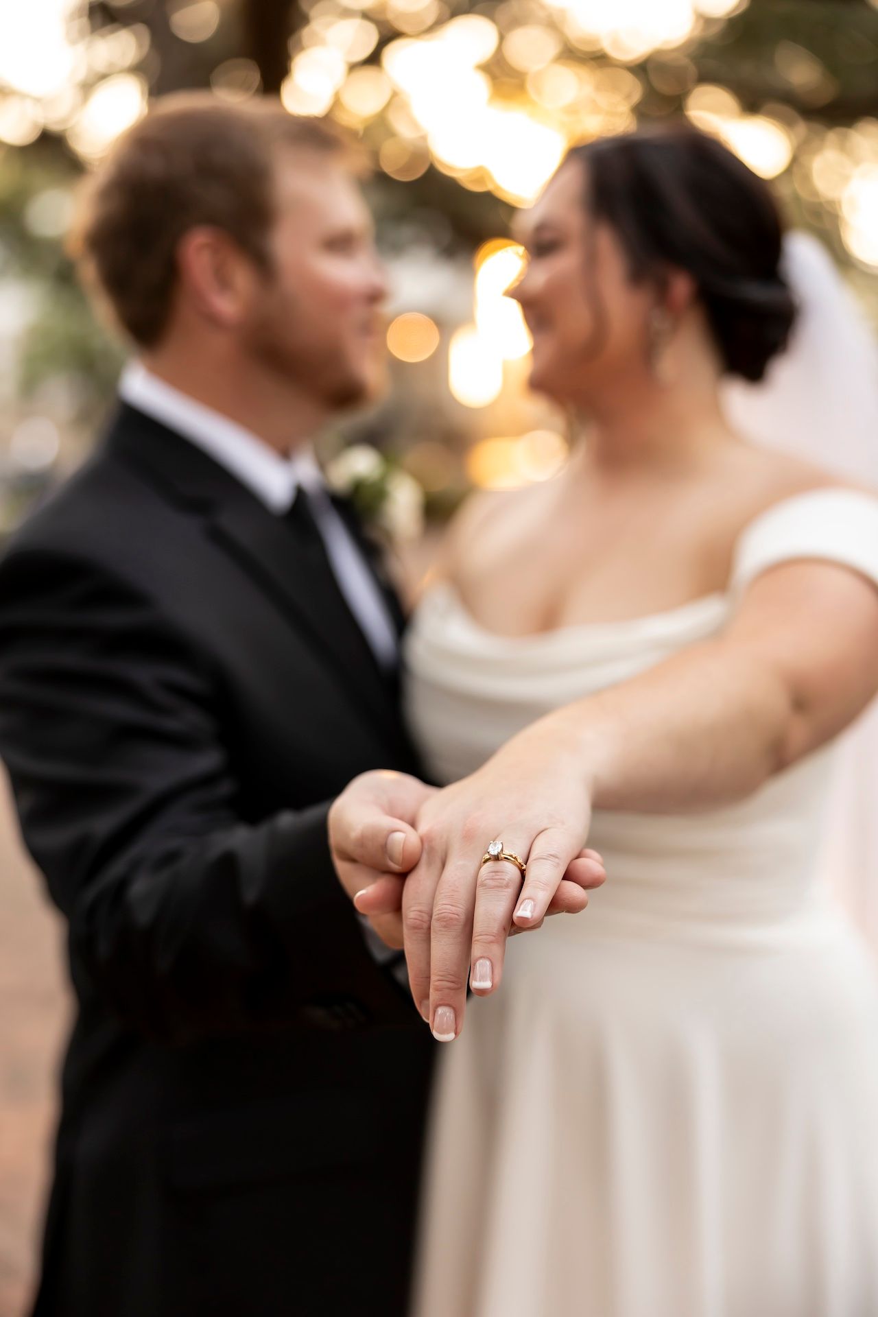 Wedding ring detail shot showing couple facing each other with soft bokeh lights in background.