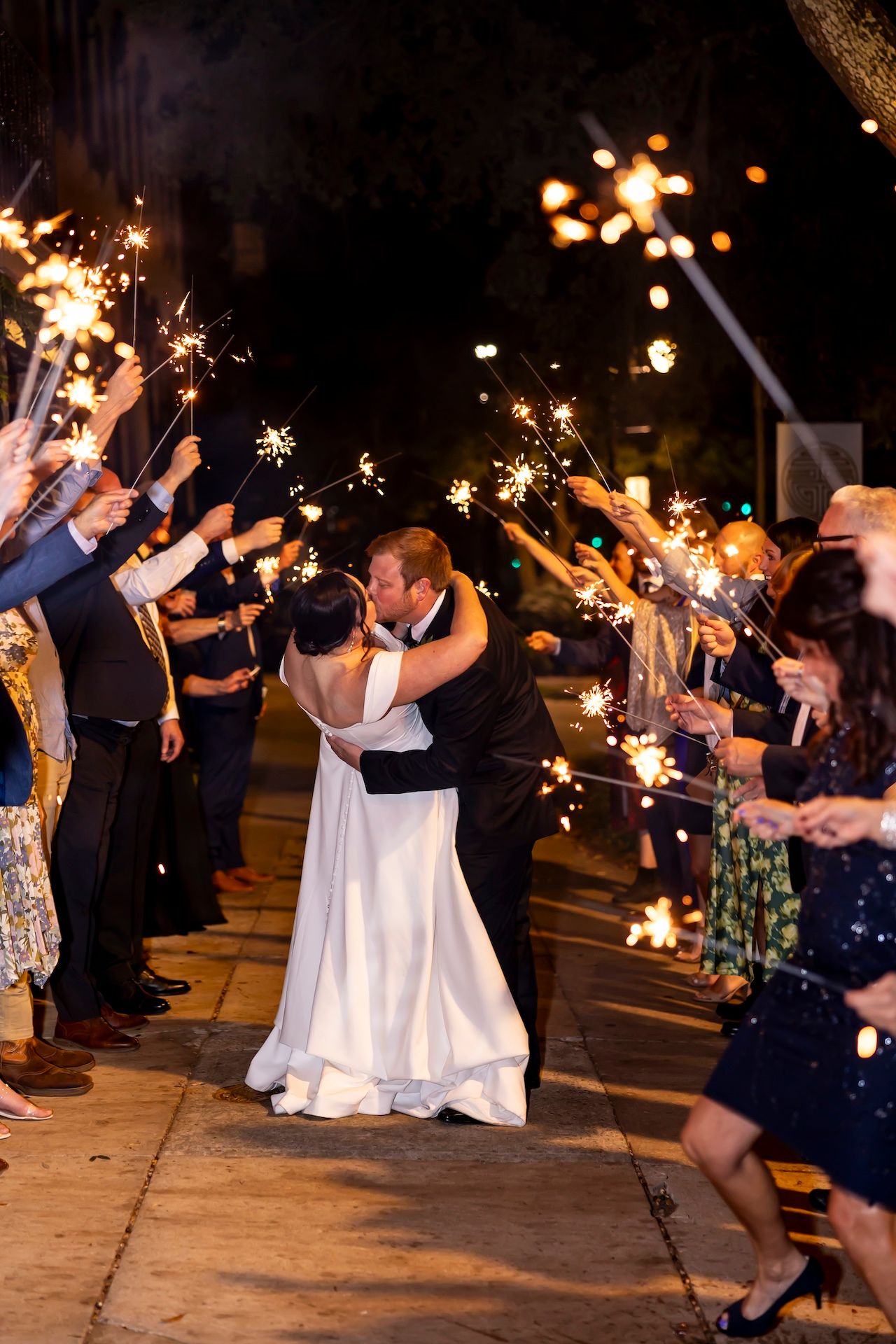 Wedding couple shares romantic kiss during sparkler send-off with guests lining pathway at nighttime celebration.
