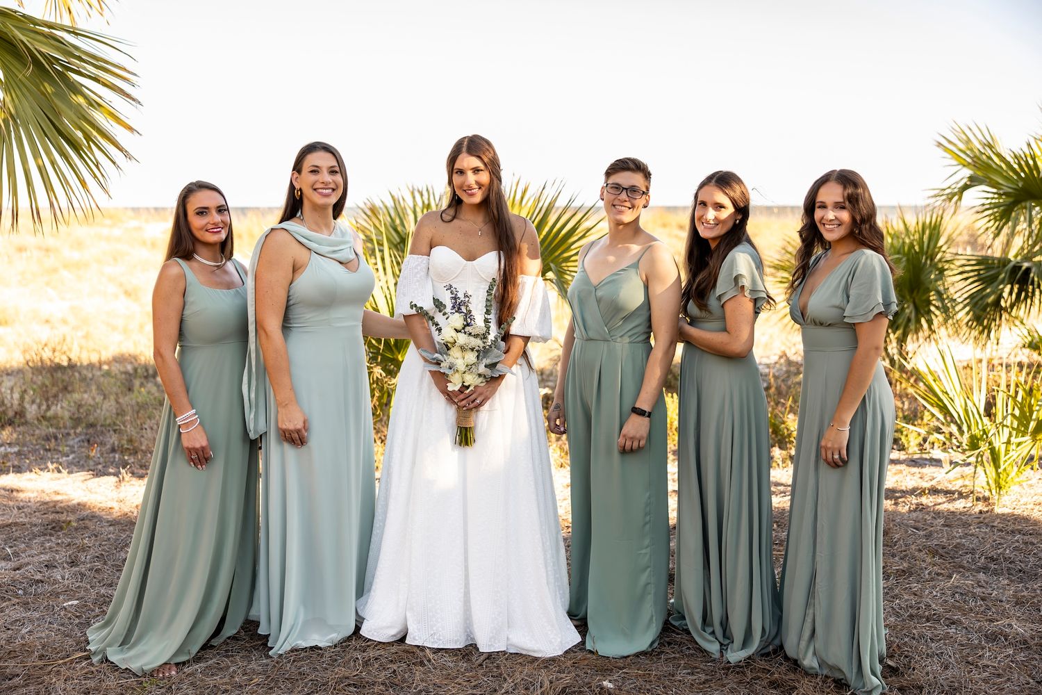 Wedding party in sage green bridesmaid dresses and white bridal gown pose together under palm trees at outdoor venue.