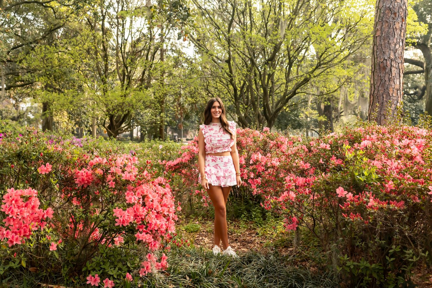 A pink floral two-piece outfit worn in a garden with vibrant red azalea bushes and trees in the background.