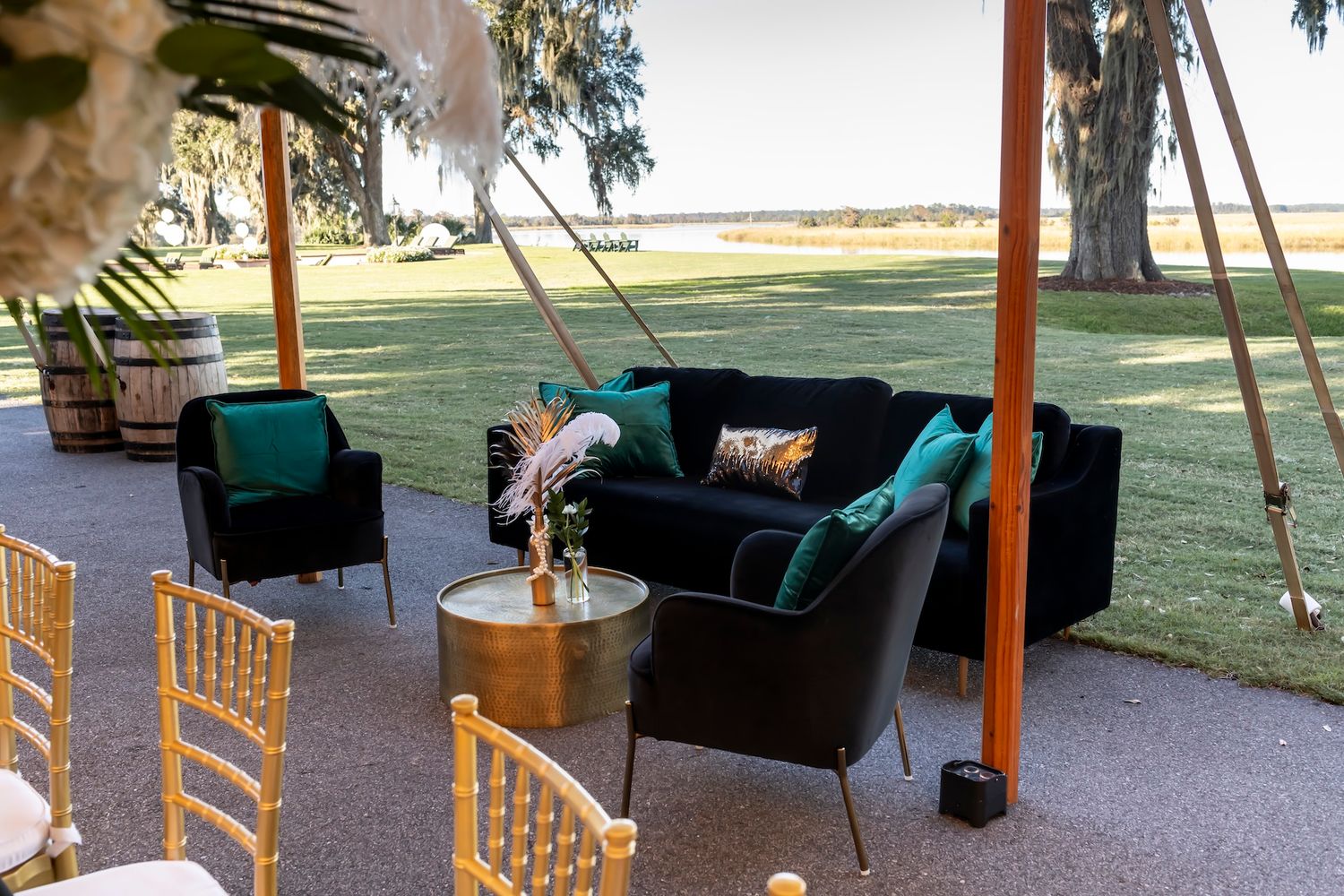 Outdoor patio lounge area with emerald green velvet sofa and gold chairs overlooking a grassy waterfront landscape.