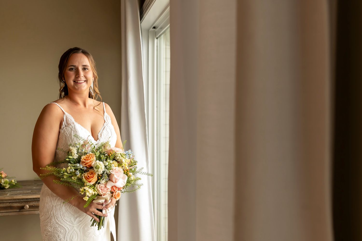 A bride in a lace wedding dress stands by a window holding a pastel bouquet with yellow and pink flowers.