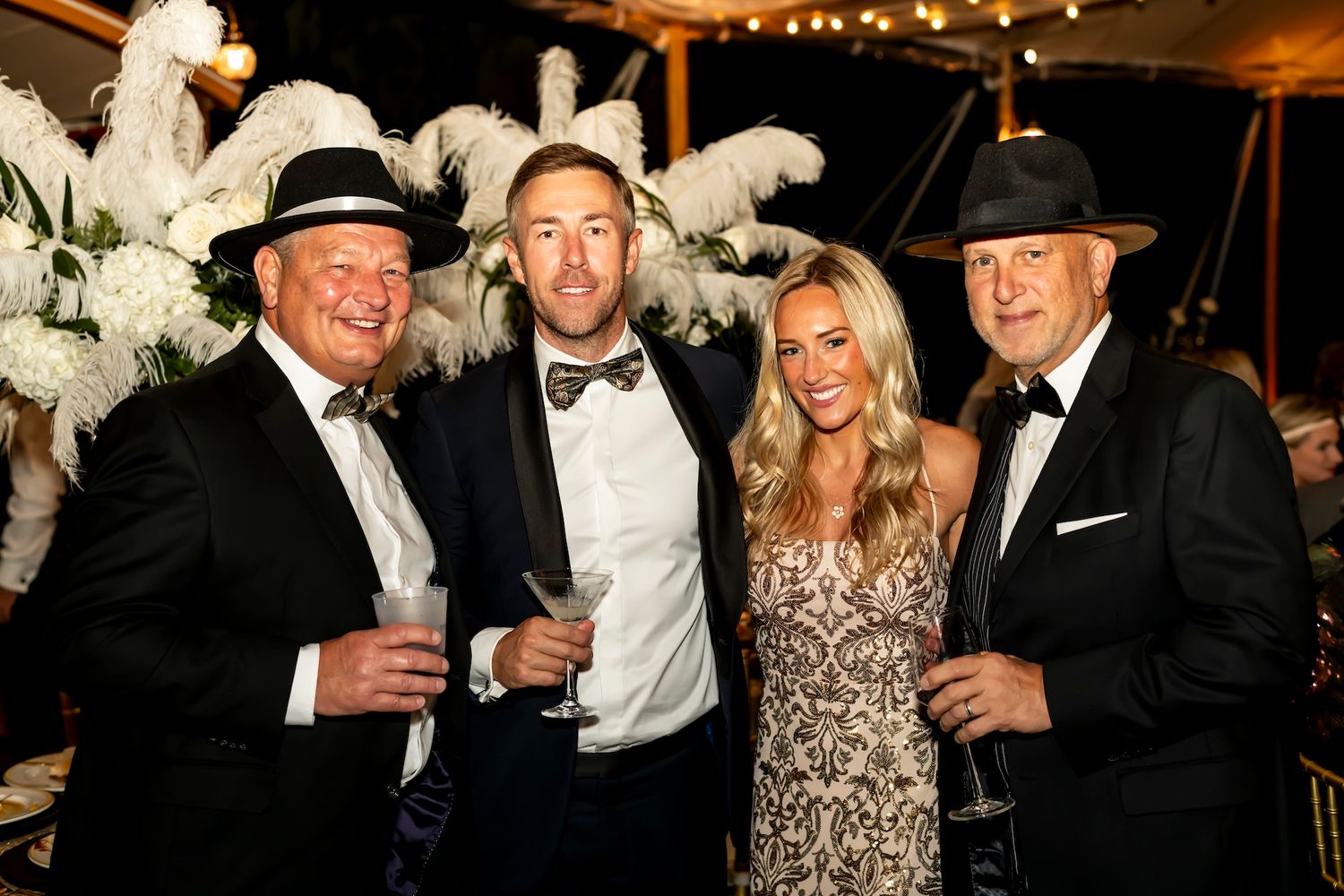 A group of formally dressed party attendees pose together with champagne glasses at an elegant event with feather decorations.