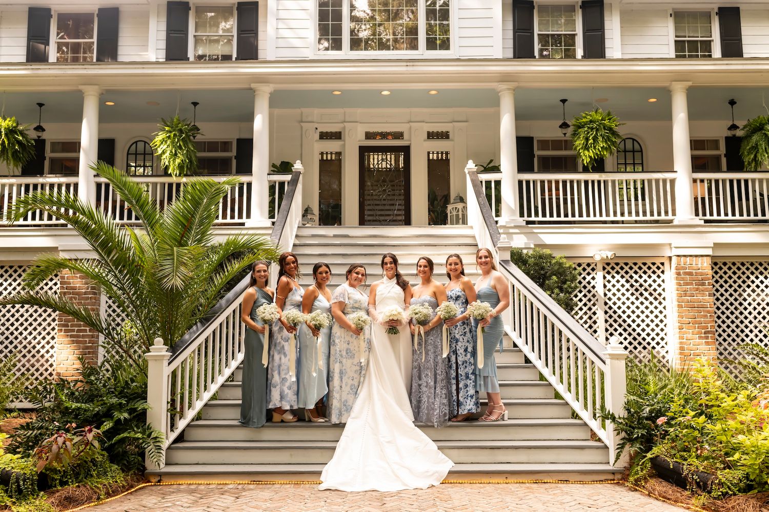 A wedding party poses on the grand staircase of a white antebellum-style mansion with a sweeping front porch and potted palms.