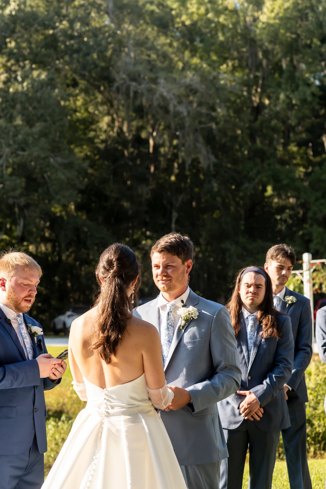 A scenic outdoor wedding ceremony takes place with the wedding party gathered under trees in golden sunlight.