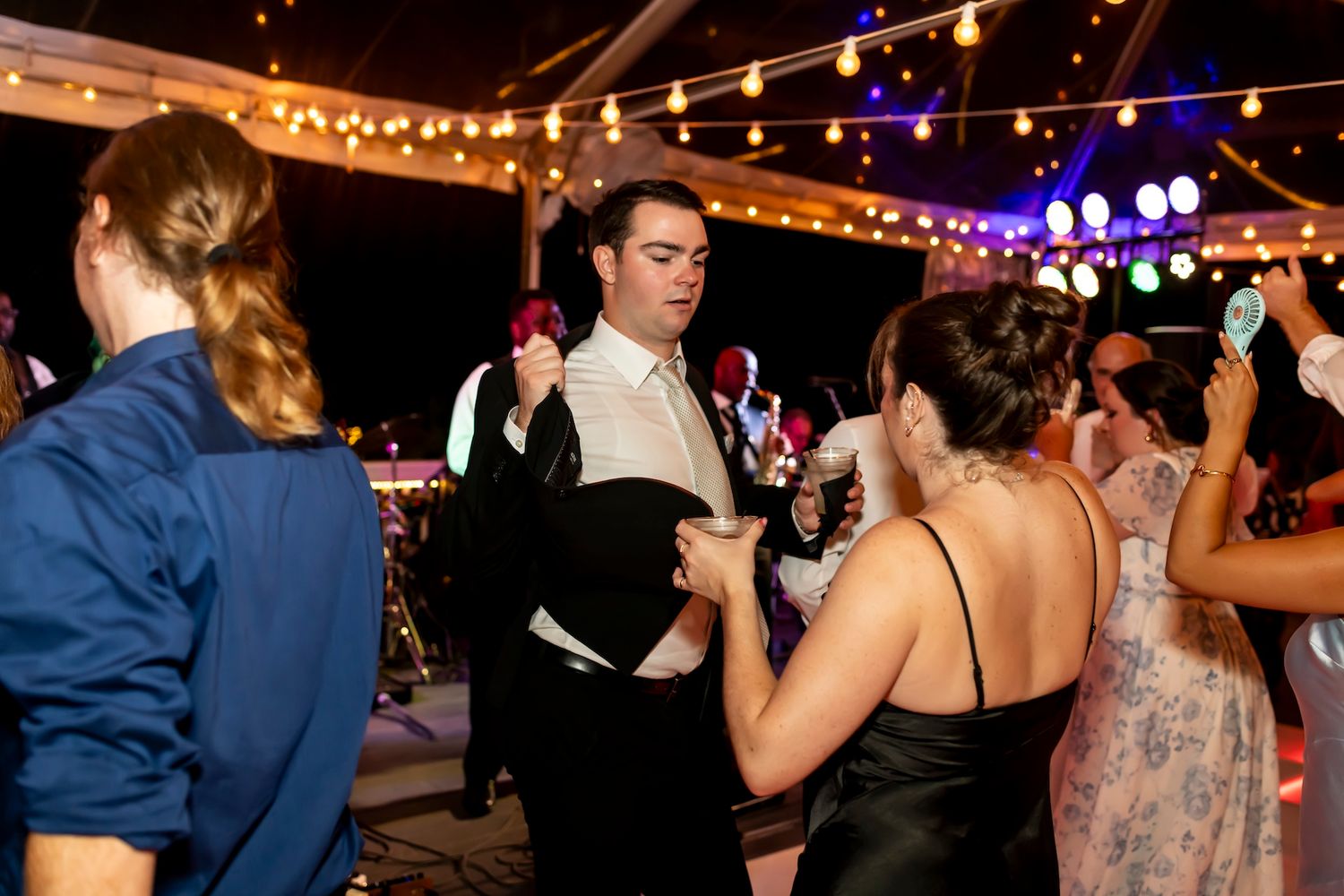 People dancing and socializing at an evening party under string lights and purple stage lighting in an indoor venue.
