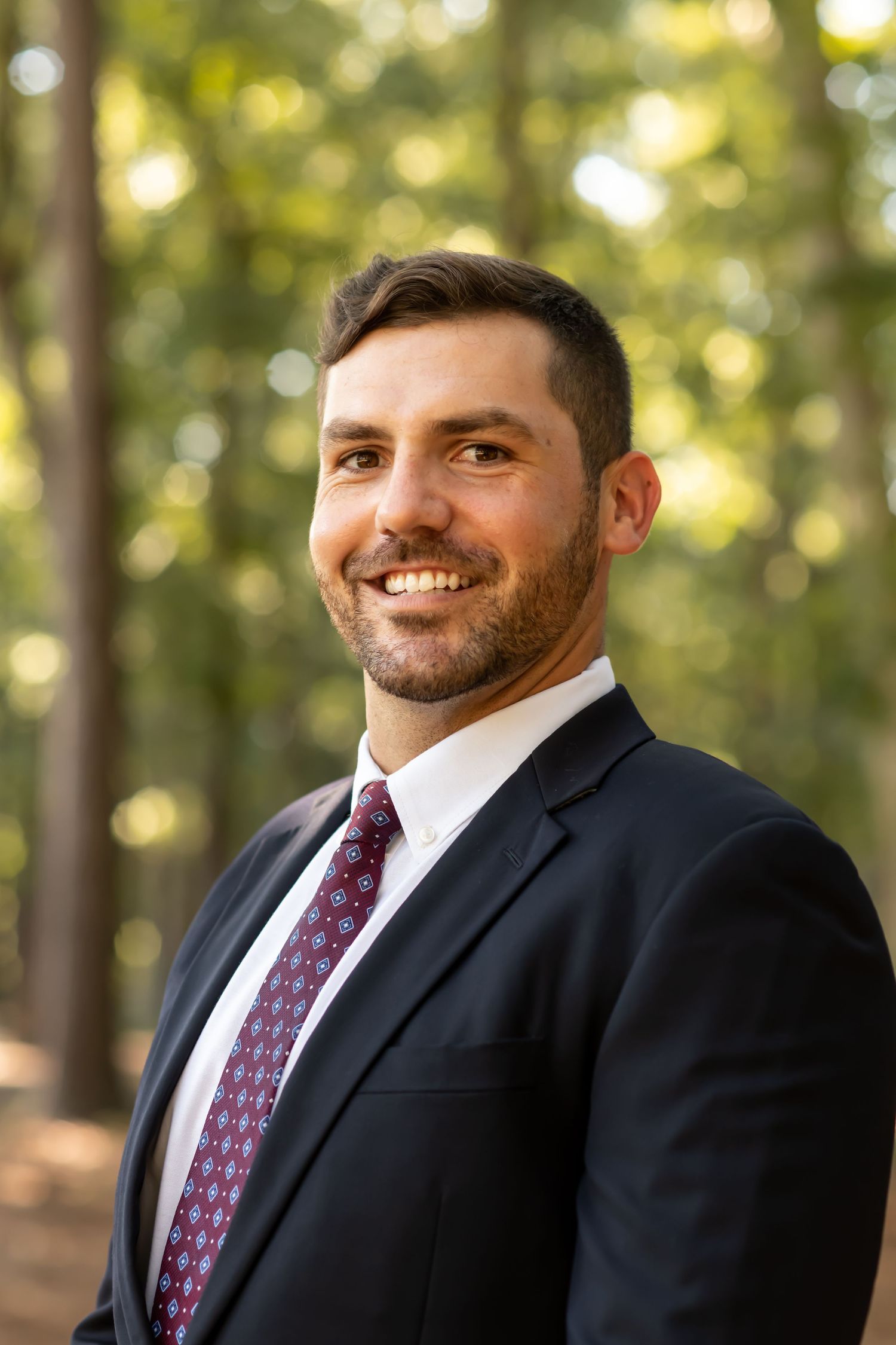 Professional headshot portraits of someone in a black suit and burgundy patterned tie against a blurred natural background.