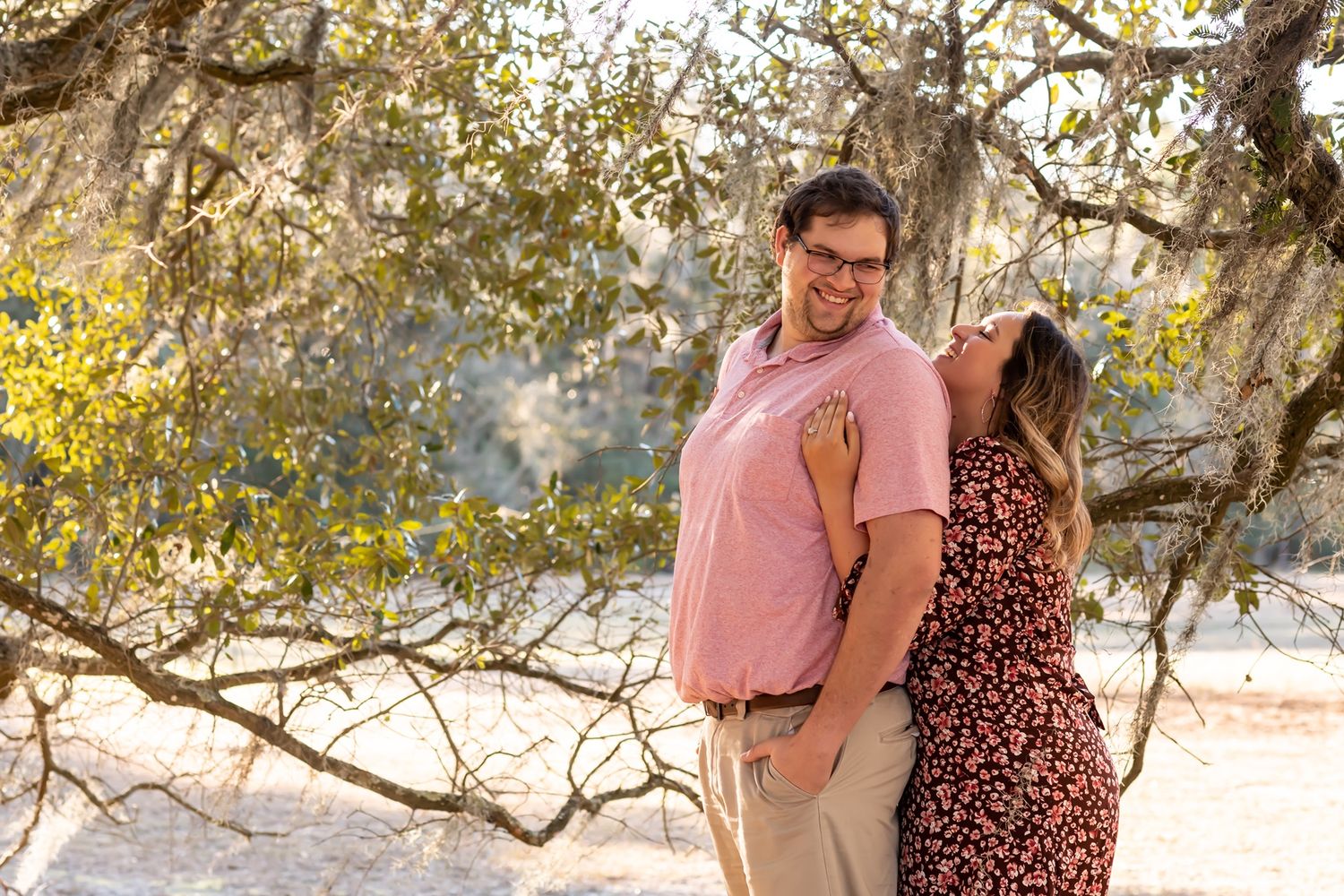 A romantic couple poses together on a sunlit wooded trail surrounded by golden autumn foliage and tree branches.