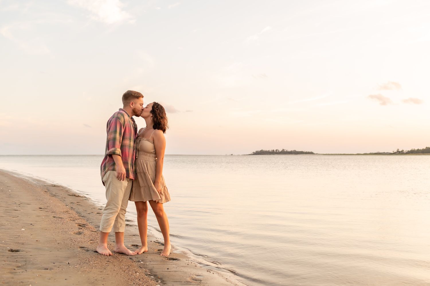 Couple strolling hand in hand along peaceful beach at sunset with calm waters and distant shoreline.