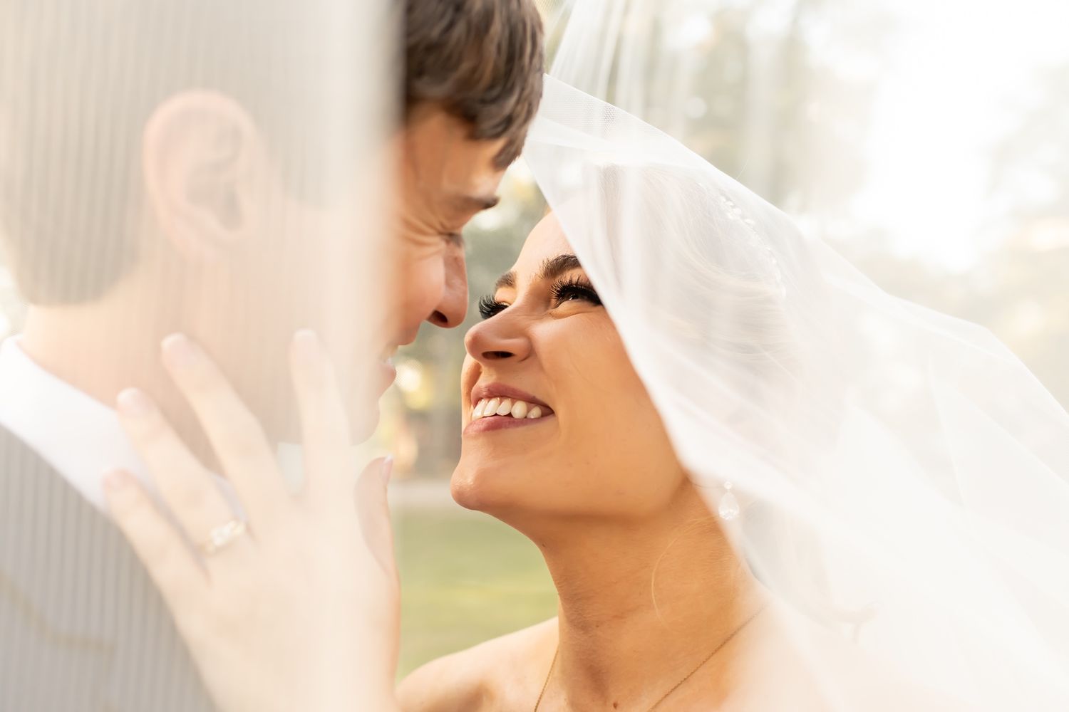 A happy newlywed couple shares an intimate moment under a wedding veil in soft natural light.
