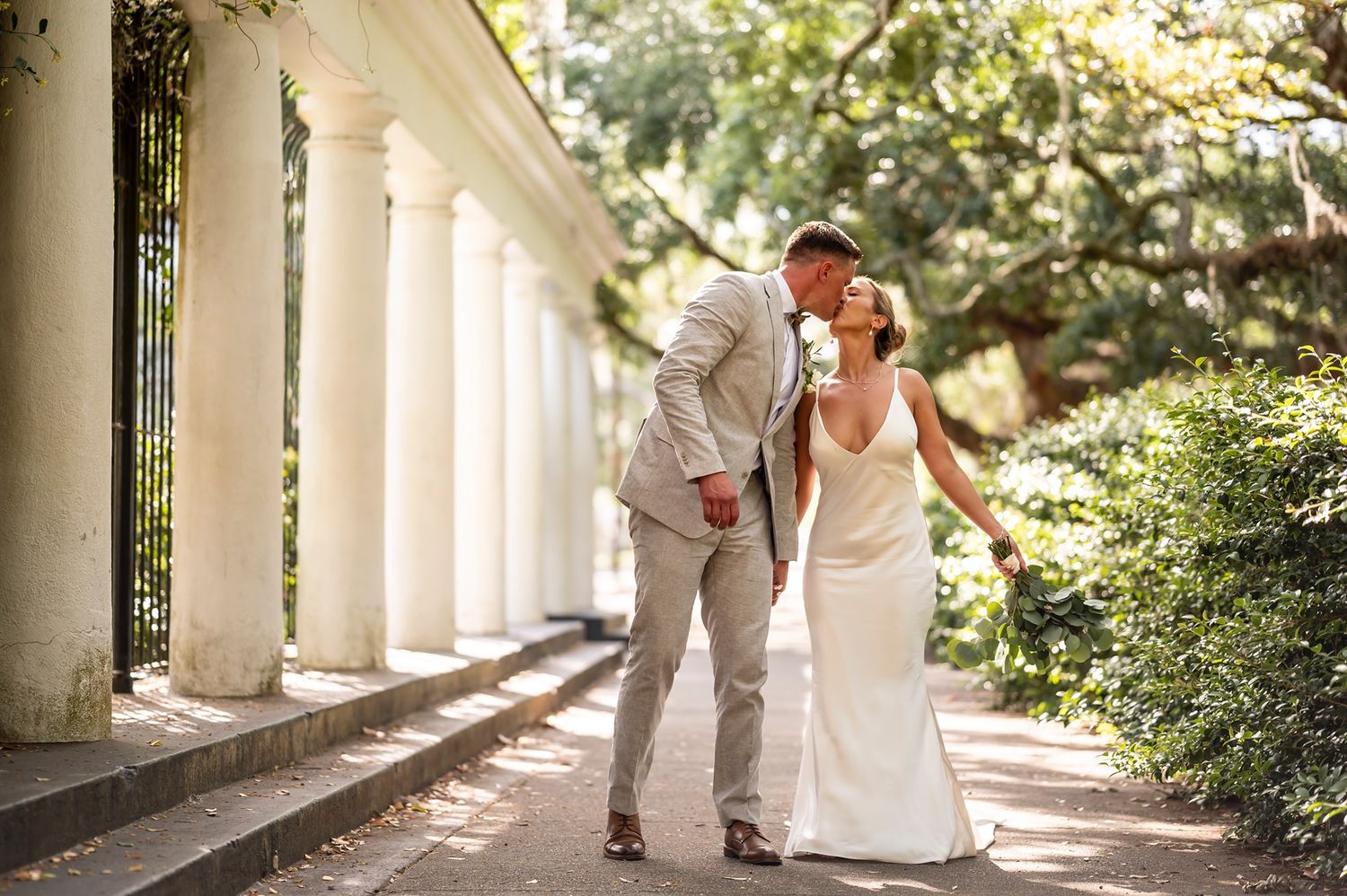 A couple in wedding attire walks together along a columned outdoor walkway on a sunny day.