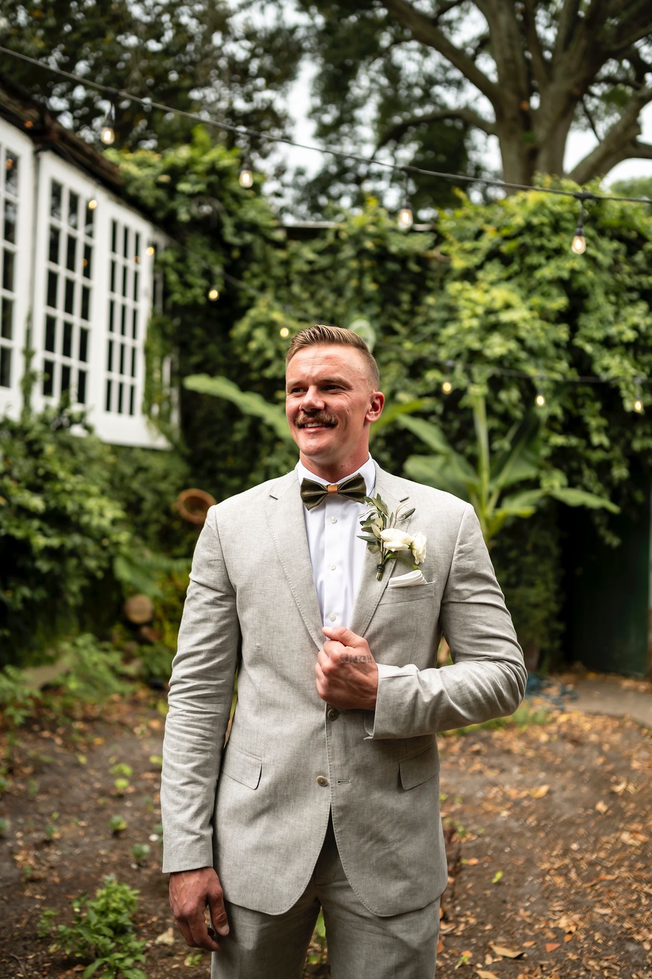 A groom in a light gray suit and bowtie stands in a garden setting on his wedding day.