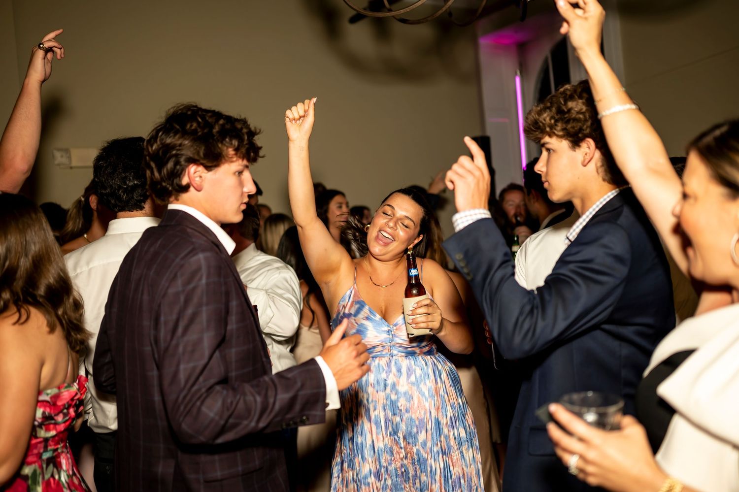Party guests in formal attire dance and celebrate with raised arms on a crowded dance floor at an indoor event.