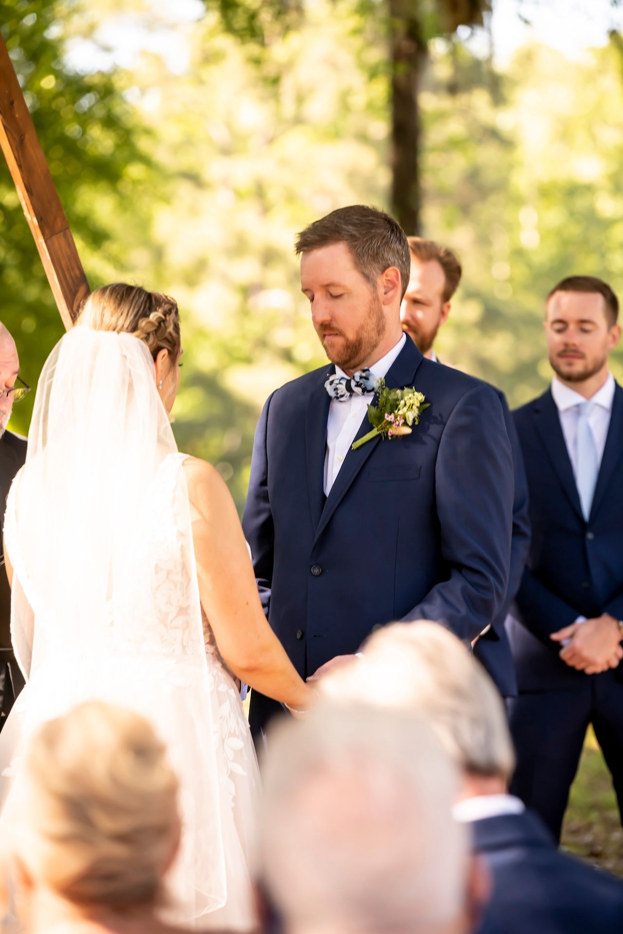 An intimate outdoor wedding ceremony takes place in a sunlit forest setting with the couple and wedding party.