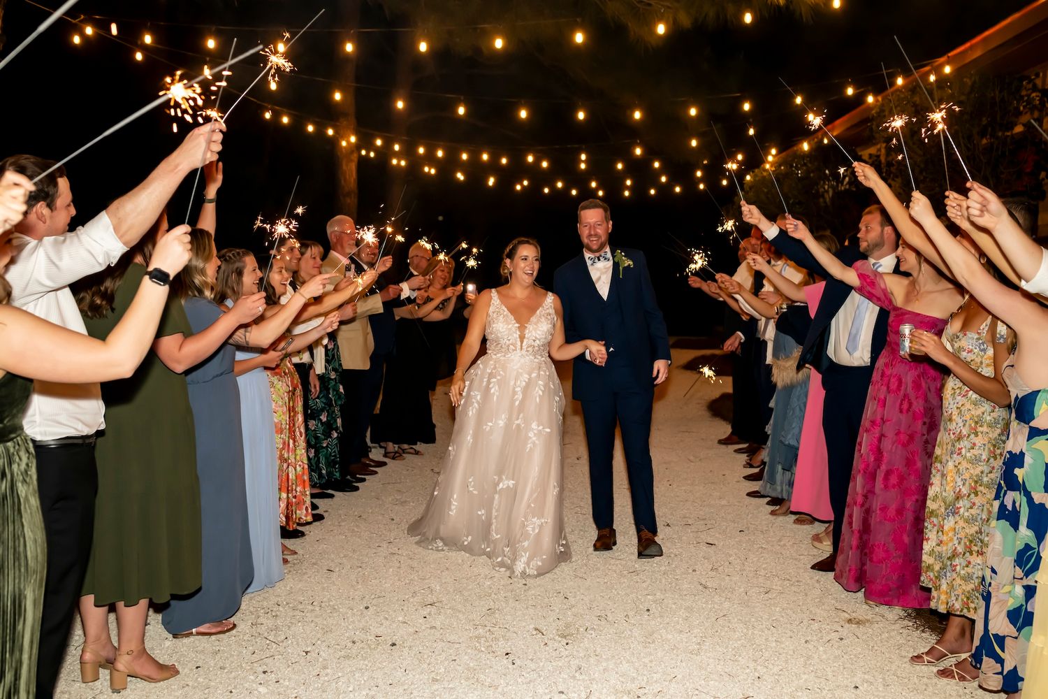 Newlyweds walk through sparkler exit line with guests in formal attire holding sparklers under string lights at night.