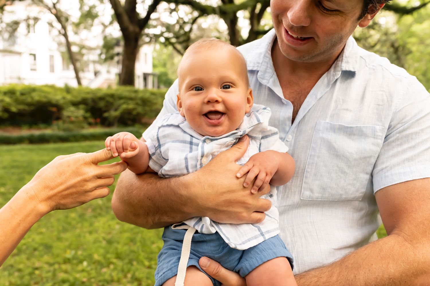 Happy smiling baby in blue and white outfit reaches toward extended hands while being held outdoors on grassy lawn.