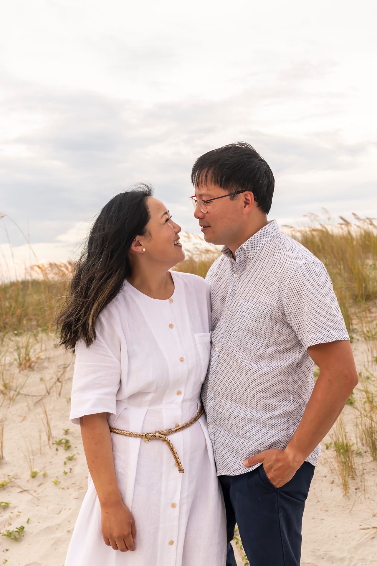 A romantic couple stands close together on sand dunes wearing white and light colored clothing at sunset.