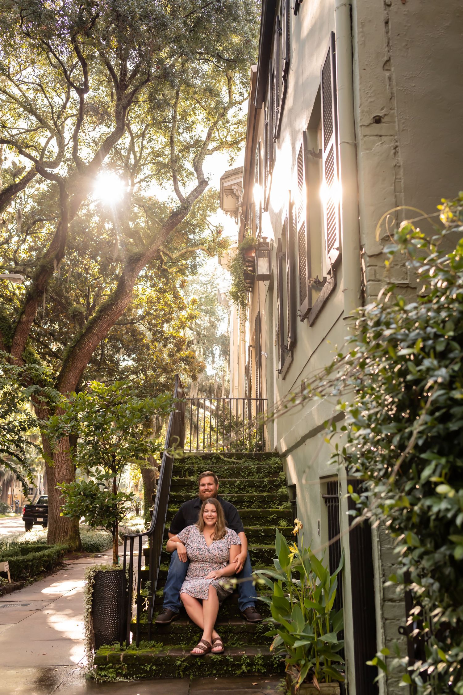 Sunlight streams through moss-draped trees beside historic Charleston steps with ornate iron railings and lush greenery.