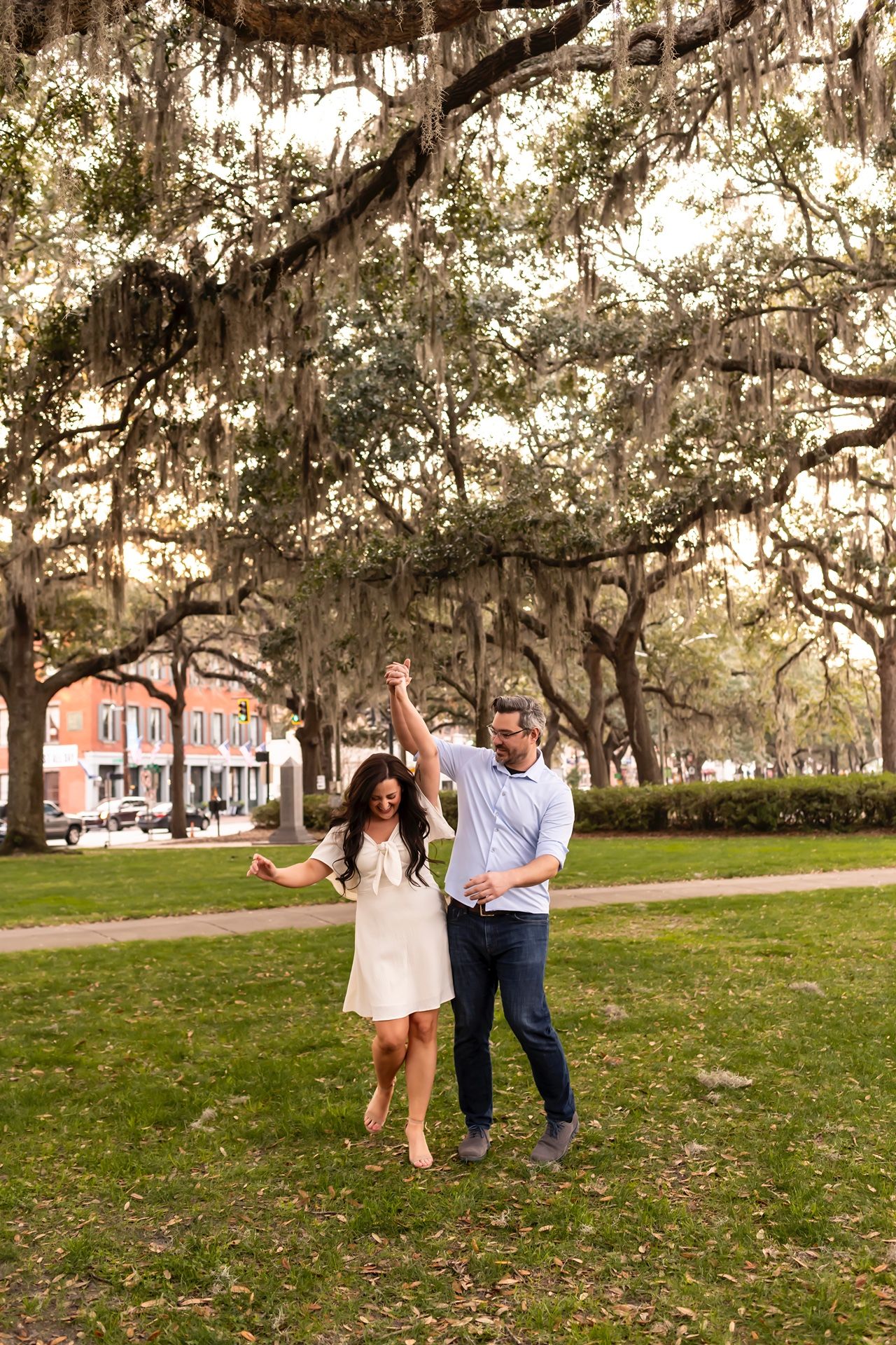 A couple dances playfully beneath Spanish moss-draped oak trees in a romantic Southern park setting during golden hour.