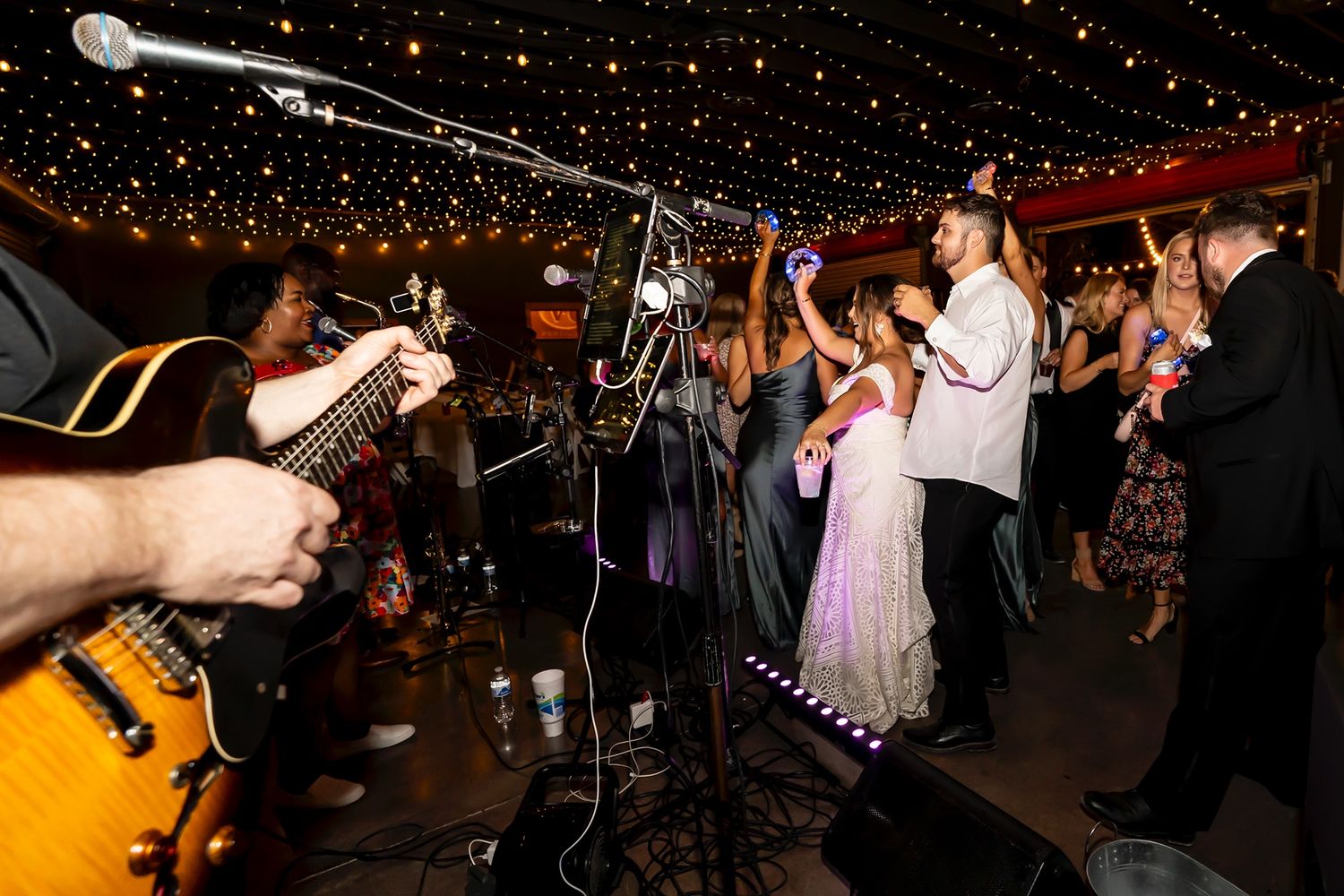 Wedding band performs as people dance and celebrate at a reception under twinkling lights.