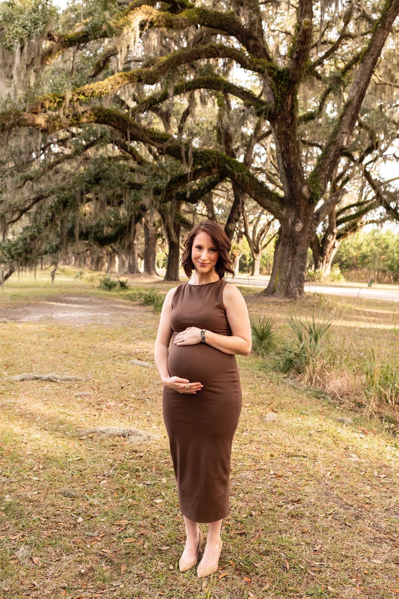 A pregnant woman in a brown dress stands under Spanish moss-draped oak trees during golden hour.