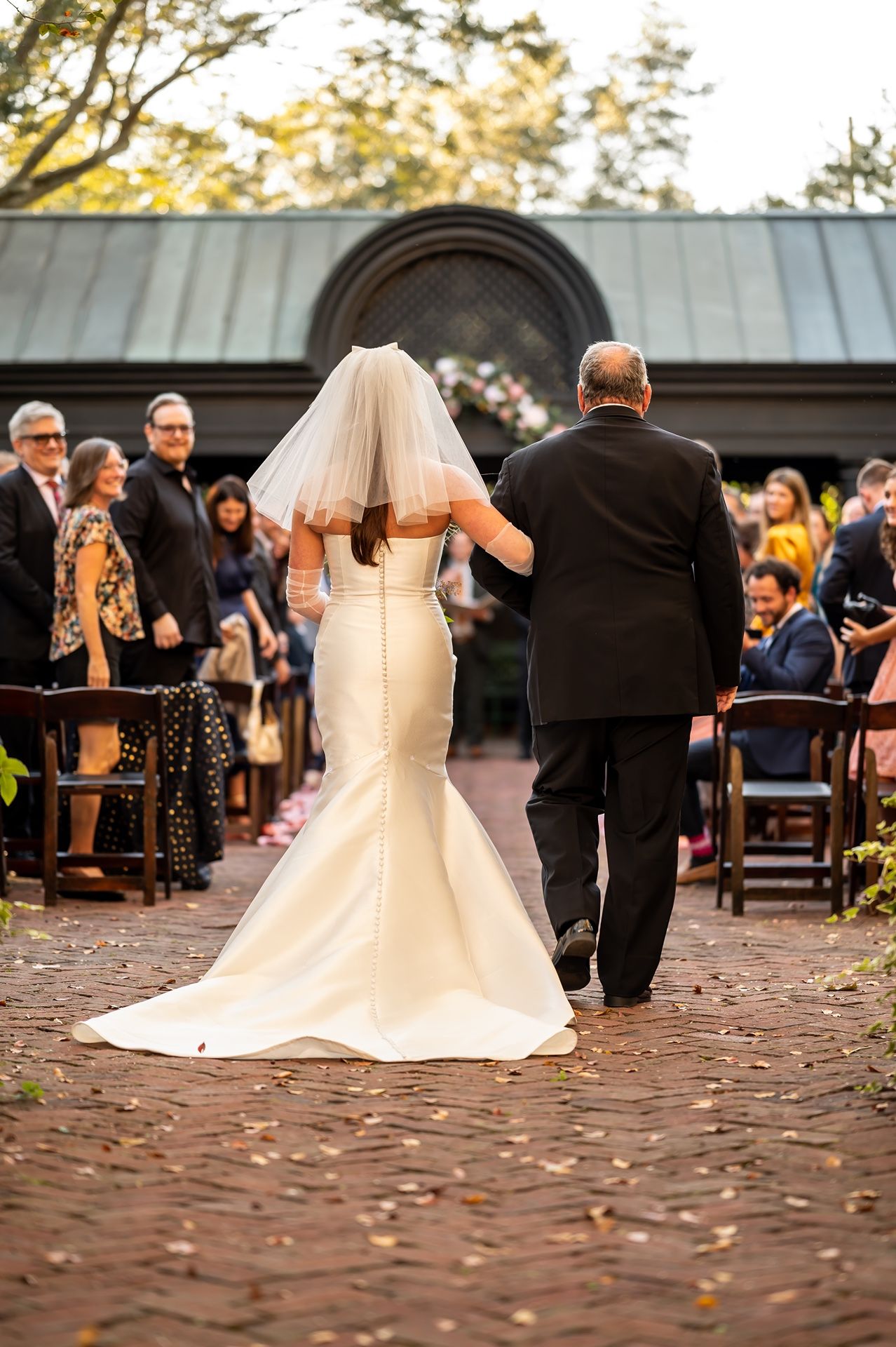 A bride in a fitted white gown and veil walks down a brick pathway with an escort at an outdoor ceremony venue.