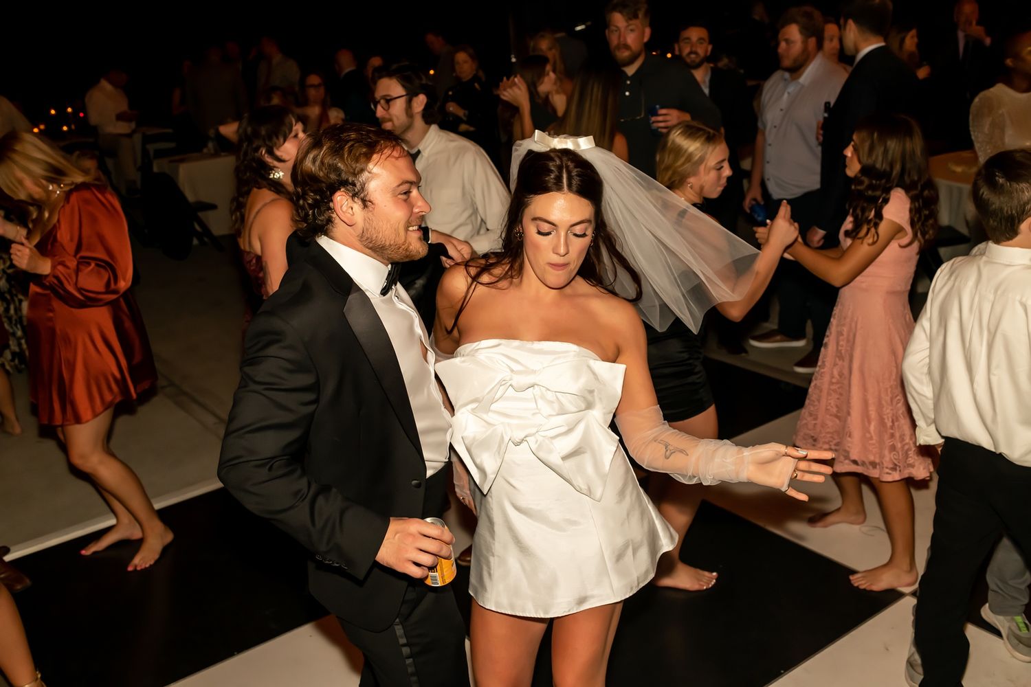 Wedding guests dance together during a reception, with guests in formal attire filling the dark dance floor.