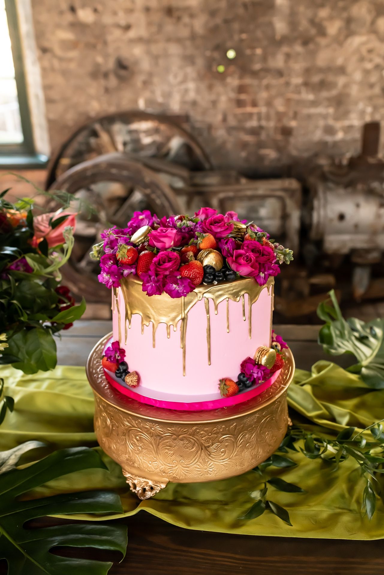 Pink drip cake decorated with vibrant fuchsia and purple flowers and fresh berries set on a gold pedestal stand.