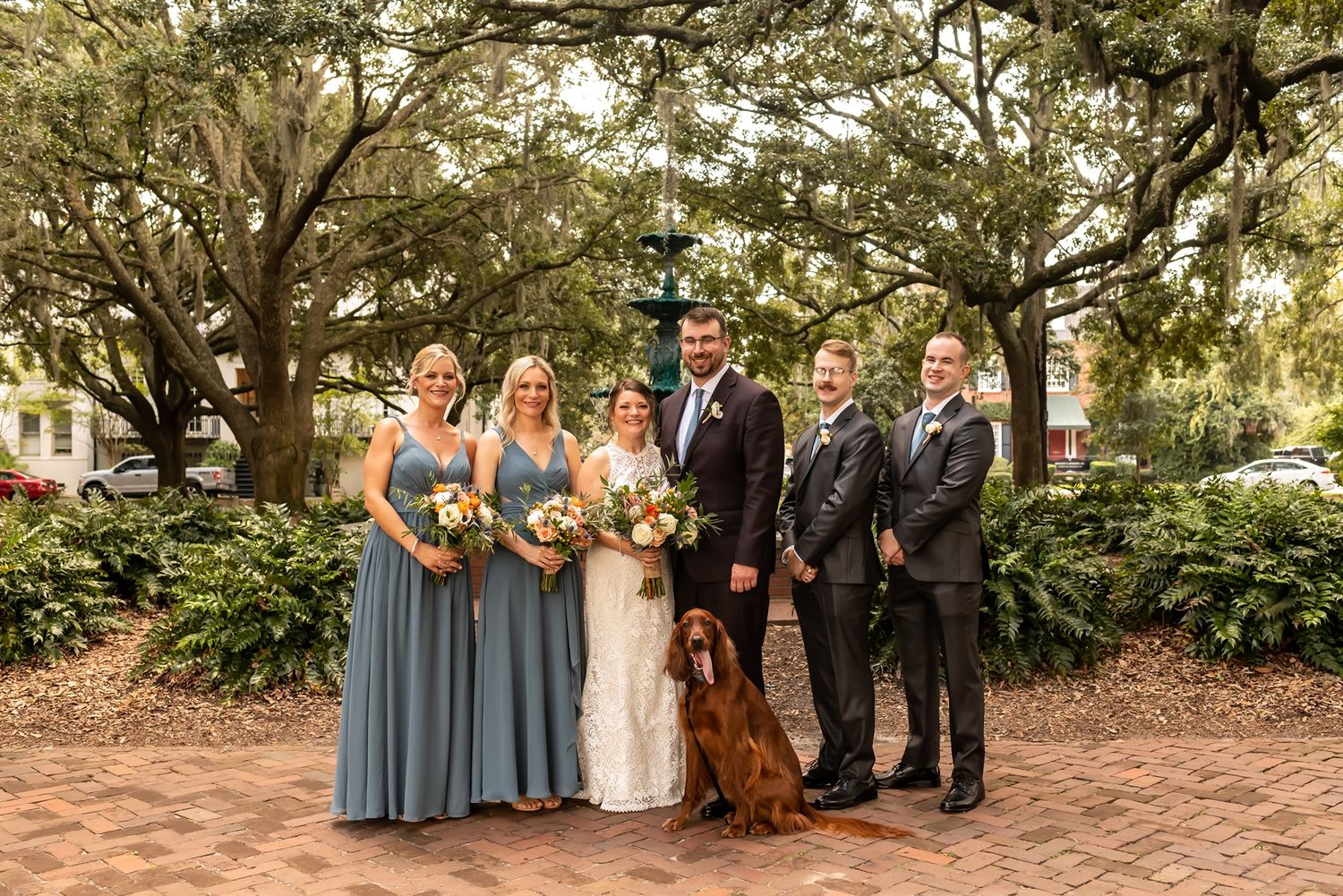 Wedding party in dusty blue dresses and gray suits poses with a brown dog in a garden setting beneath oak trees.