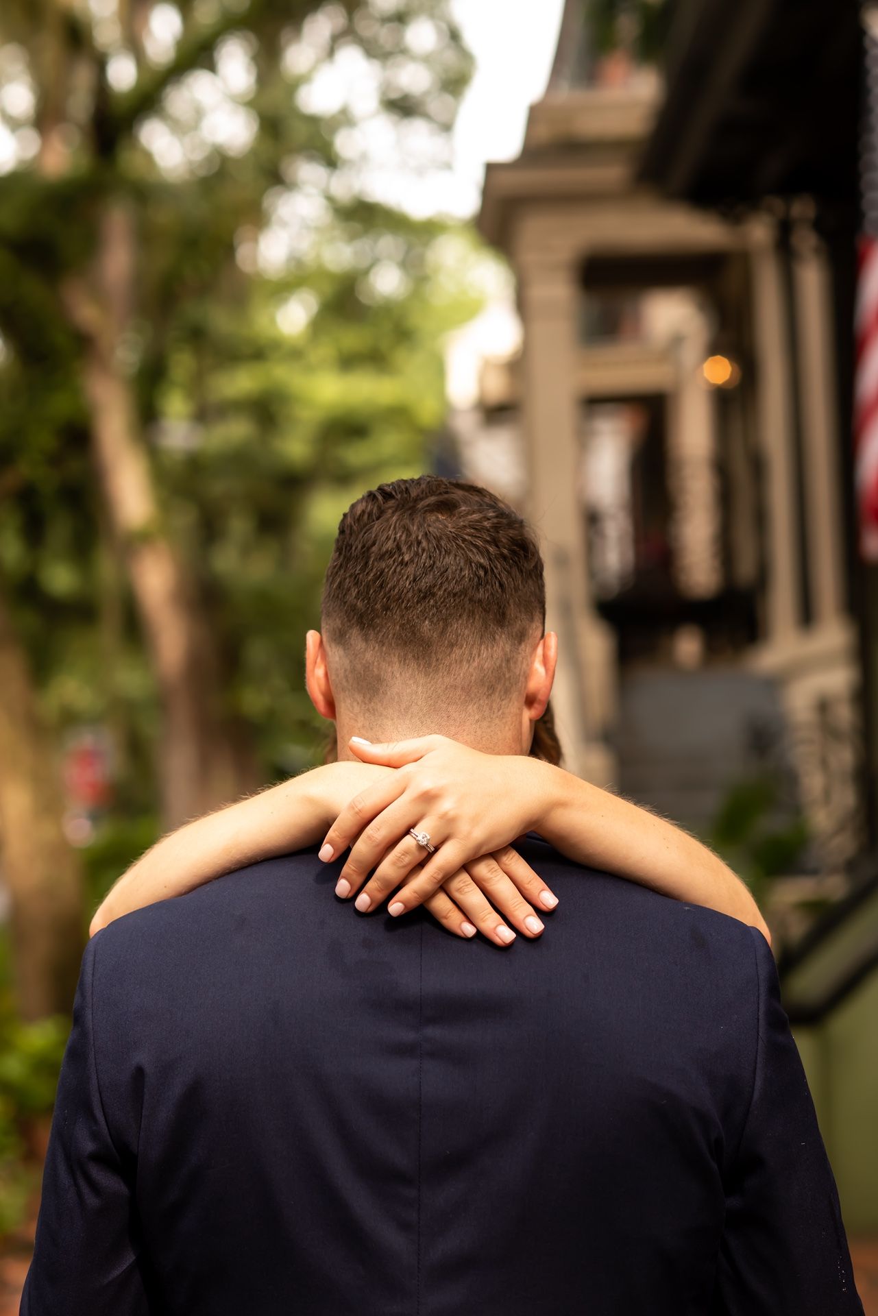 Back view of person in navy suit with arms embraced around their shoulders in front of blurred urban building exterior.