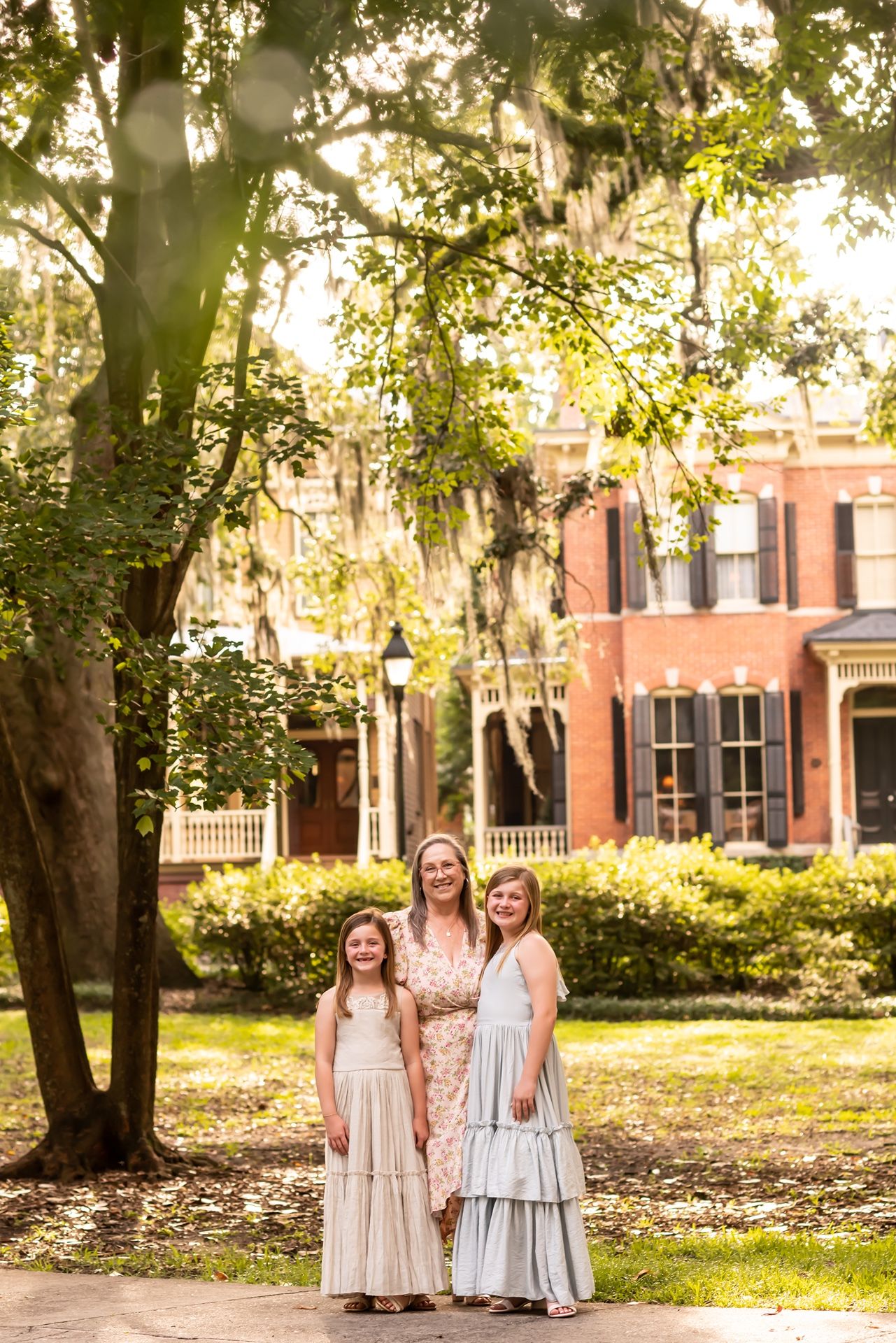 Golden sunlit garden scene with Spanish moss draped trees in front of a historic brick mansion in the South.