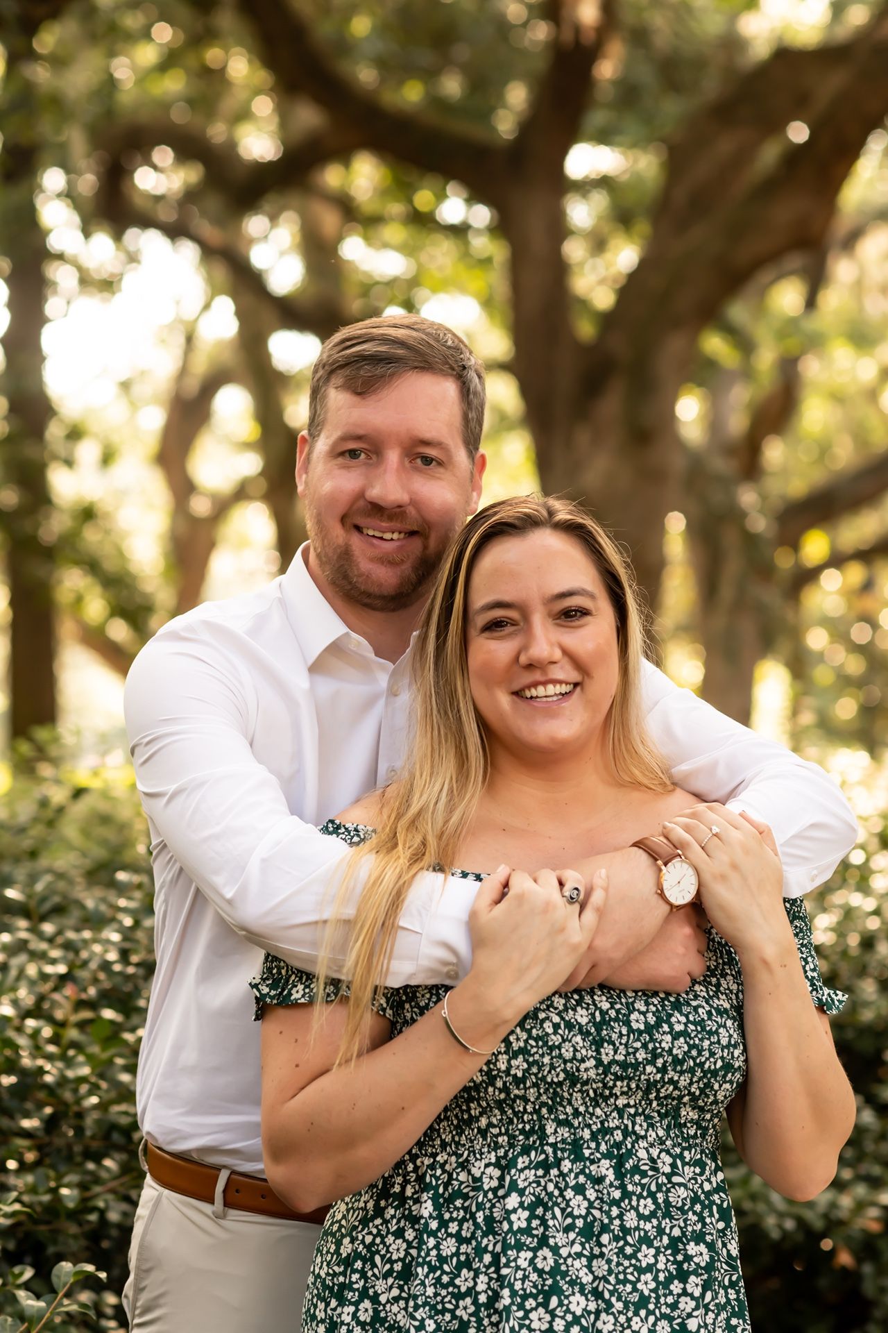 A couple embraces in a romantic outdoor portrait with sunlit trees and greenery in the background.