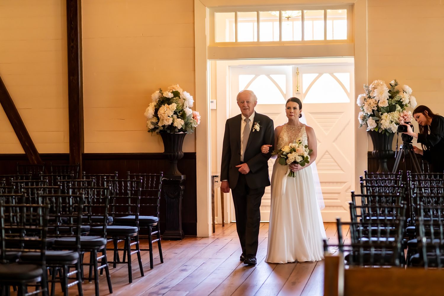 A glowing ceremony venue with an elegant aisle lined with black chairs and floral arrangements at the entrance doorway.